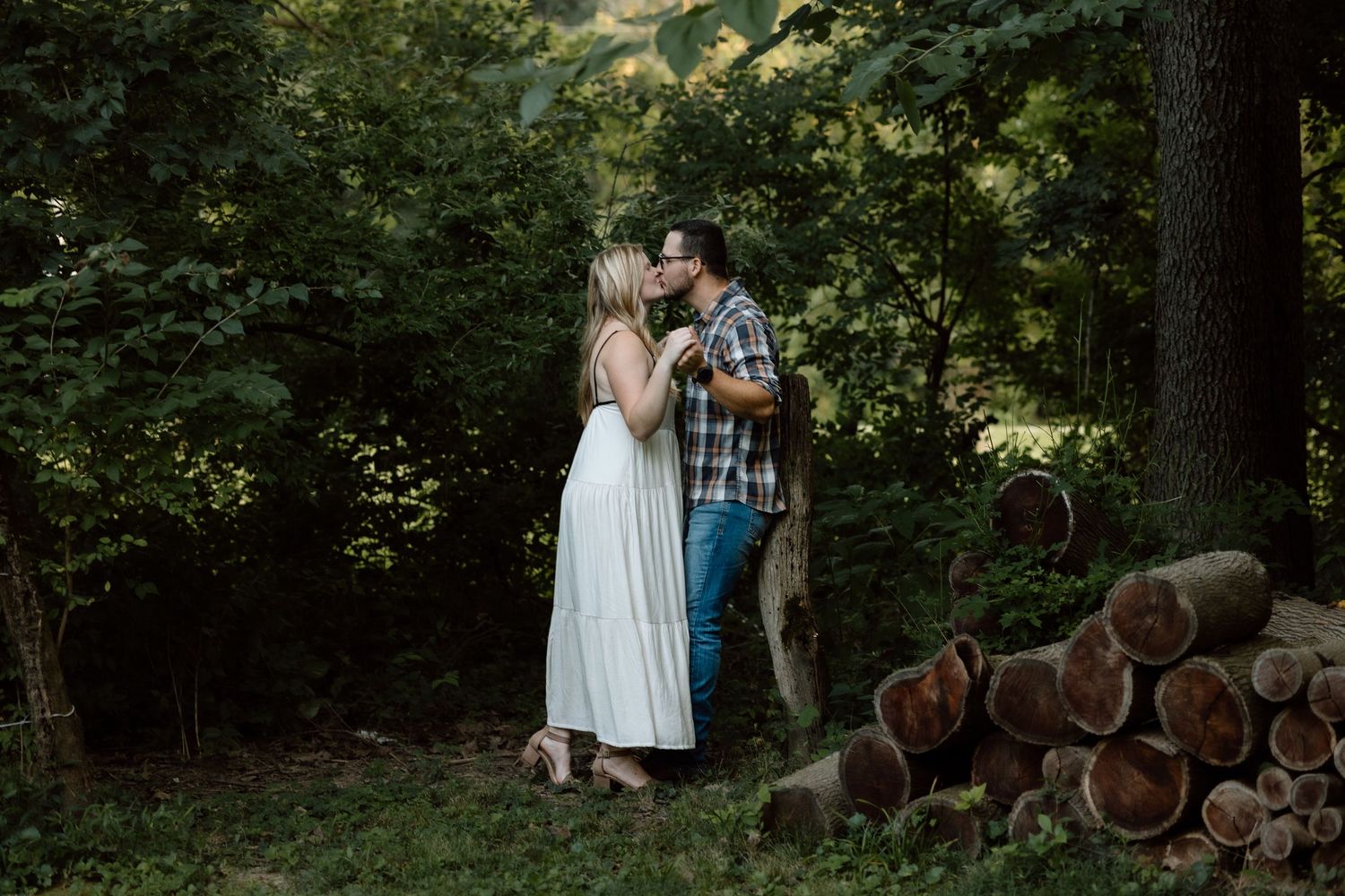 An engaged couple shares a kiss in a wooded area with stacked logs in the background during a photo shoot.