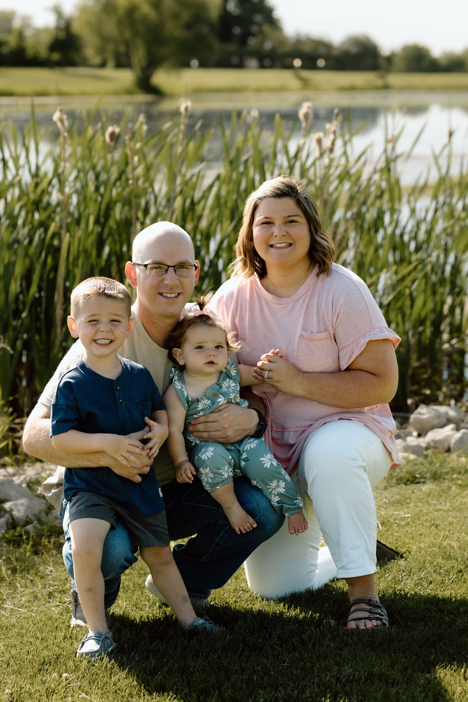 Family of four smiles together near a pond, parents posing with young son and baby girl on grass.