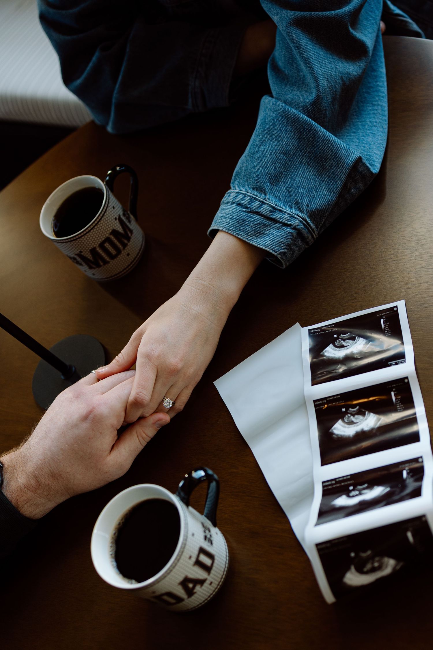 Overhead view of couple holding hands near Mom and Dad mugs with ultrasound photos on table.