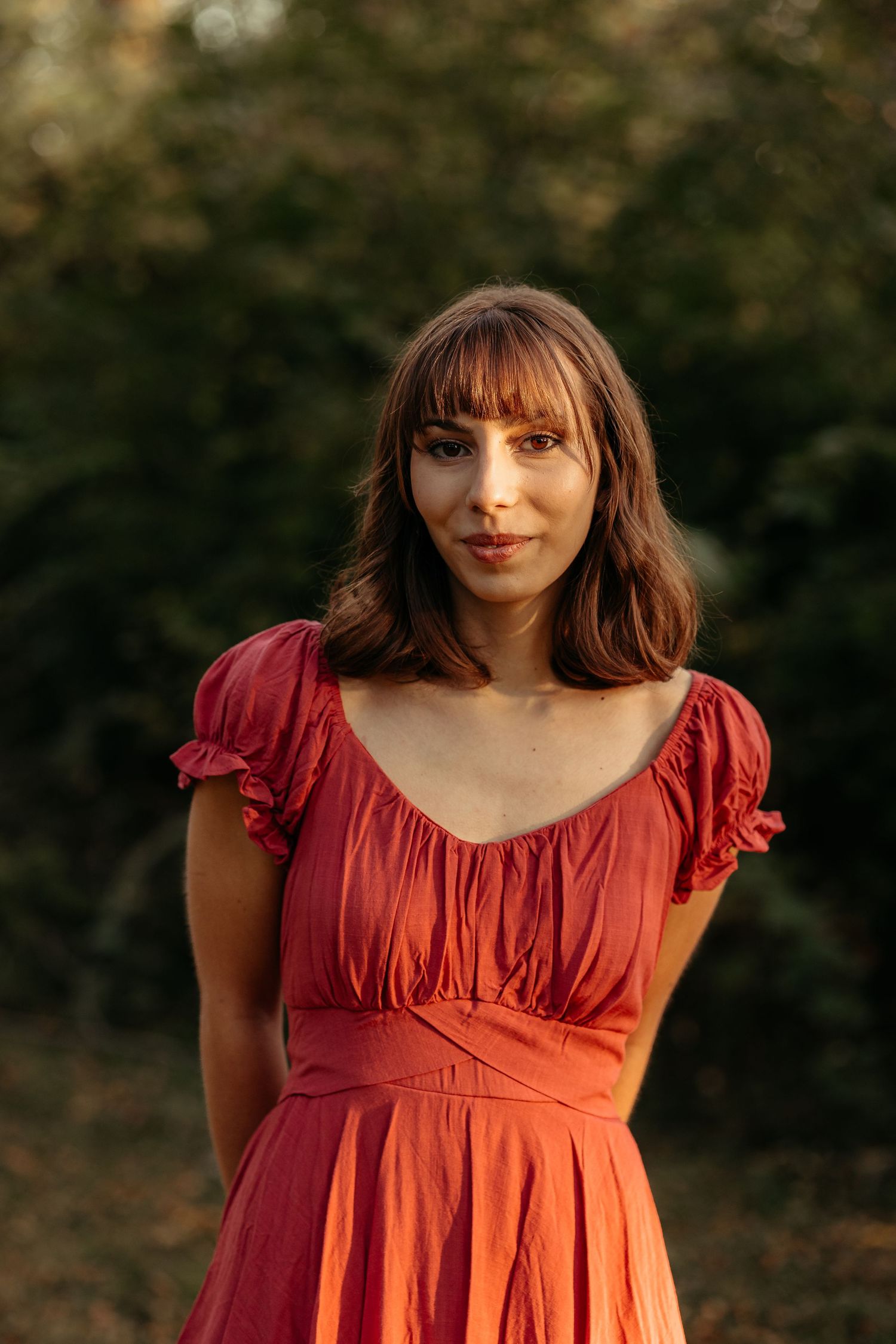 Young woman in red dress standing outdoors with lush green trees in background.