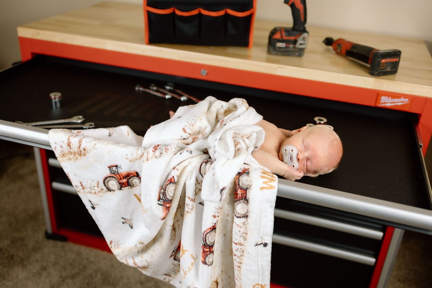 Sleeping newborn baby wrapped in tractor-print swaddle blanket lying on a Milwaukee tool workbench.