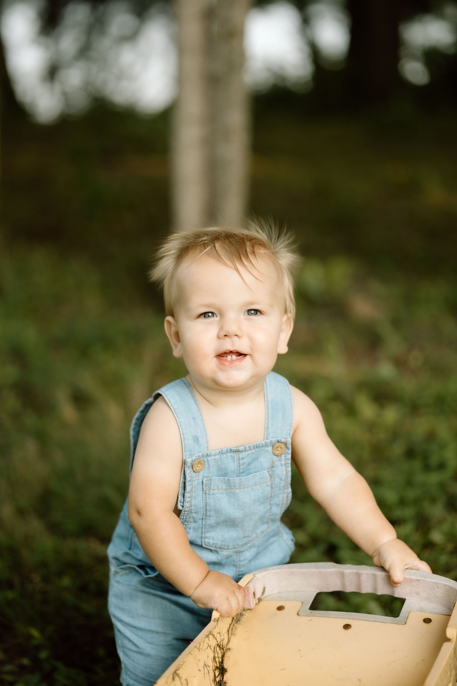 Happy toddler in blue denim overalls smiling while holding wooden toy outside.