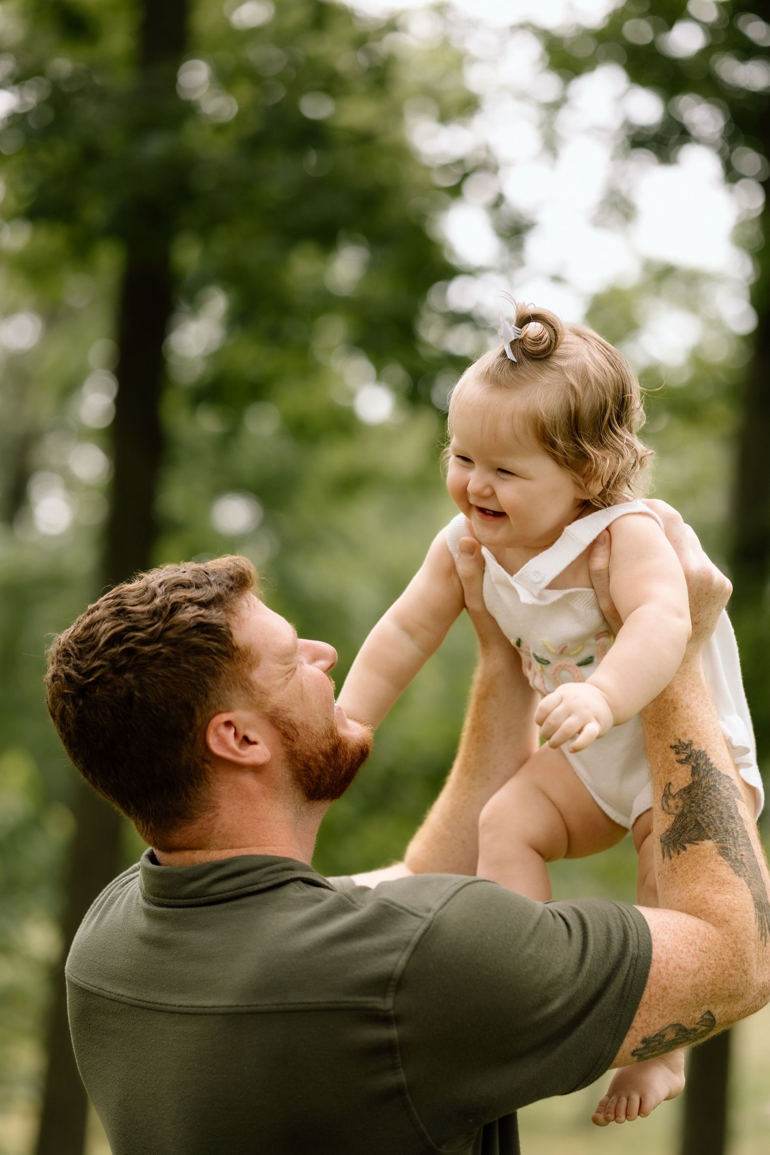 Tattooed father lifts laughing baby girl in the air outdoors in a green park.