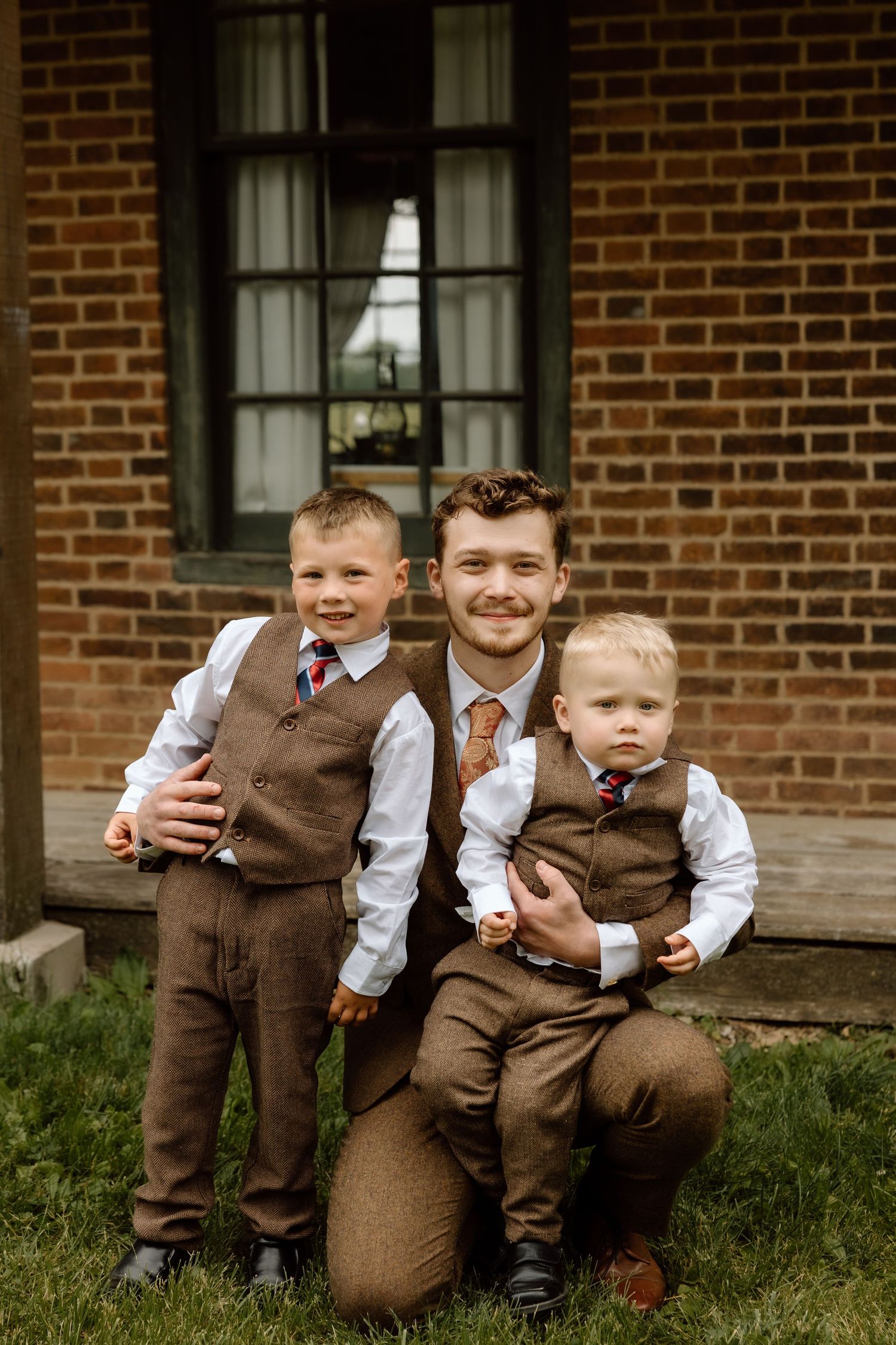 Groom in brown tweed vest crouching with two young ring bearers in matching brown vests before brick wall.