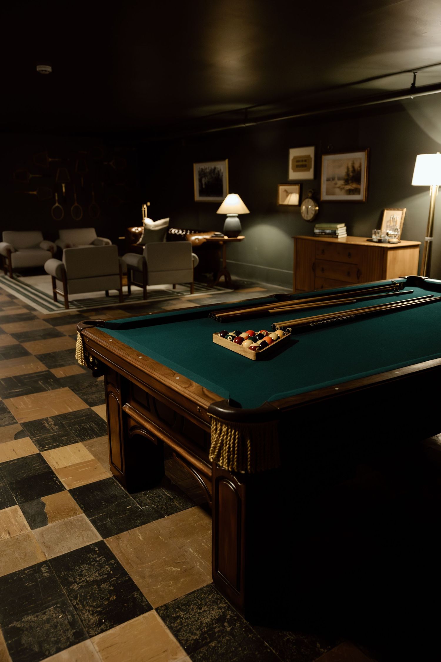 Dimly lit billiard room with green felt pool table, cue sticks, and balls on checkered tile floor with seating area.