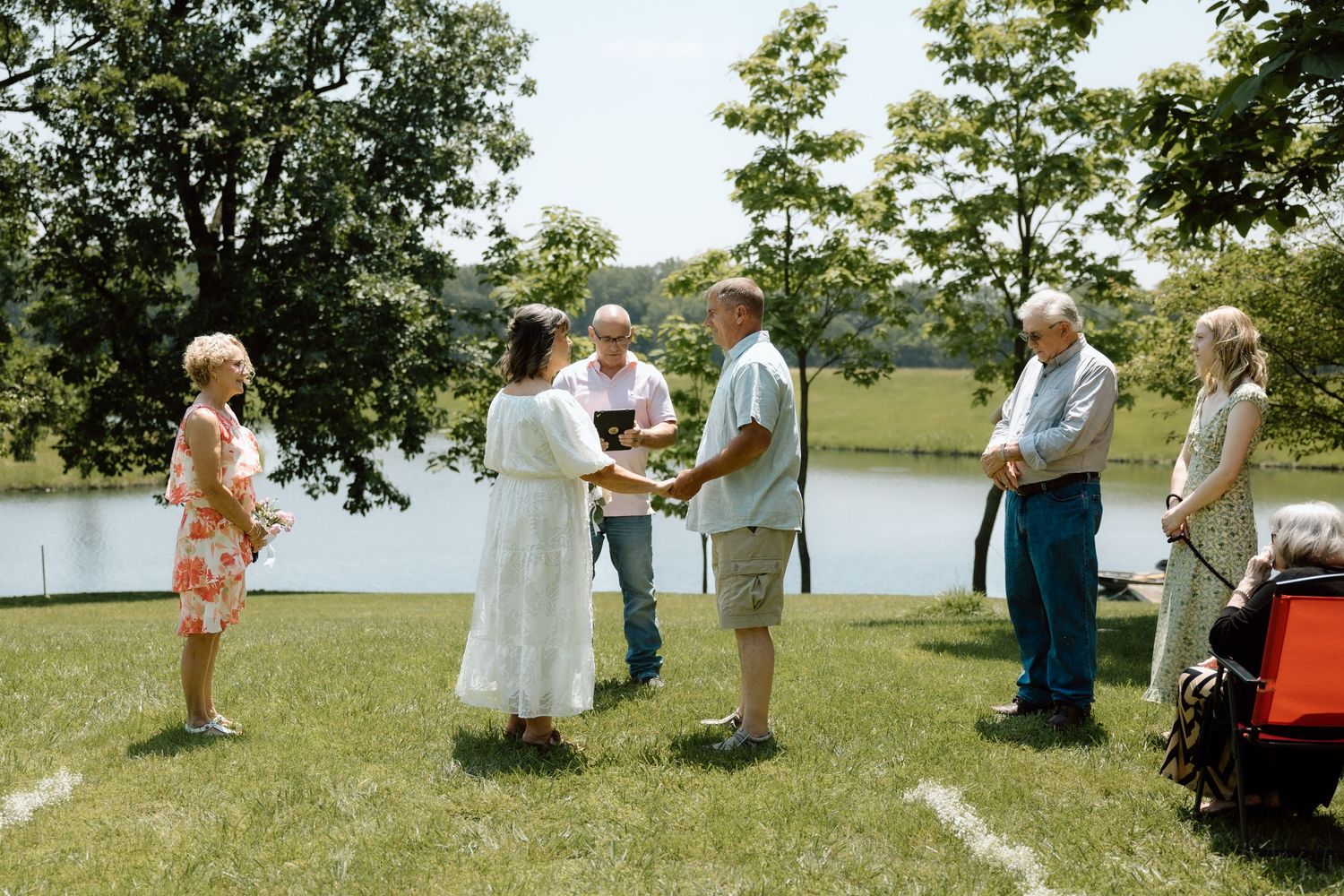 Intimate outdoor wedding ceremony beside a serene lake with small group of guests on a sunny day.