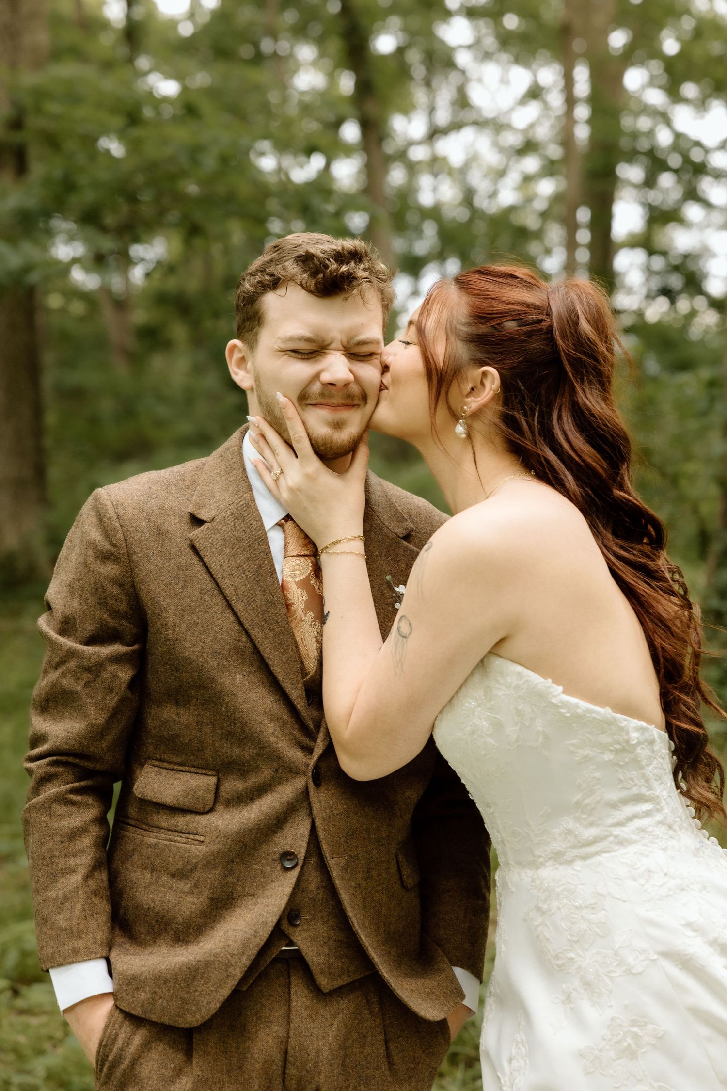 Bride kisses groom on cheek outdoors, groom smiling in brown tweed suit with floral tie.