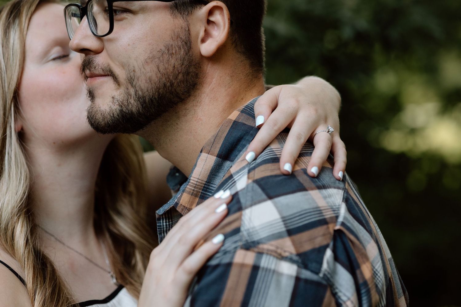 A woman with an engagement ring kisses a bearded man on the cheek during a romantic outdoor photo session.