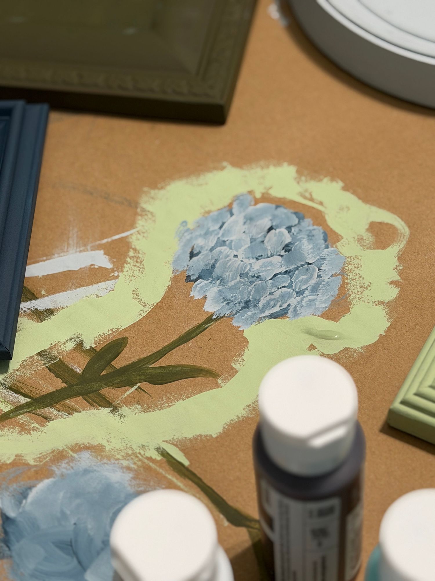 Blue hydrangea flower painting in progress on kraft paper with acrylic paint bottles nearby.