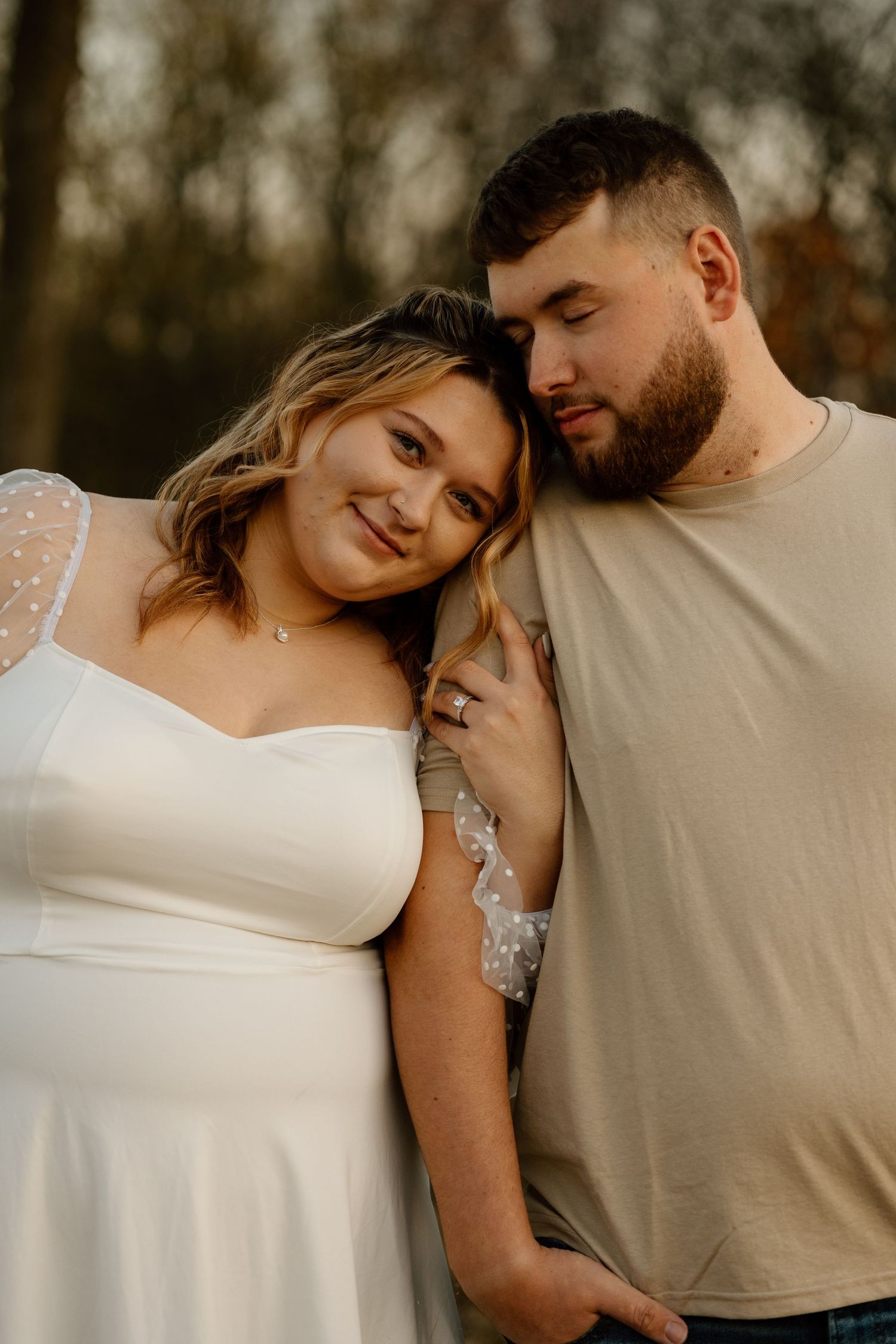 Couple sharing an intimate moment outdoors, woman in white lace dress leaning against bearded man at golden hour.