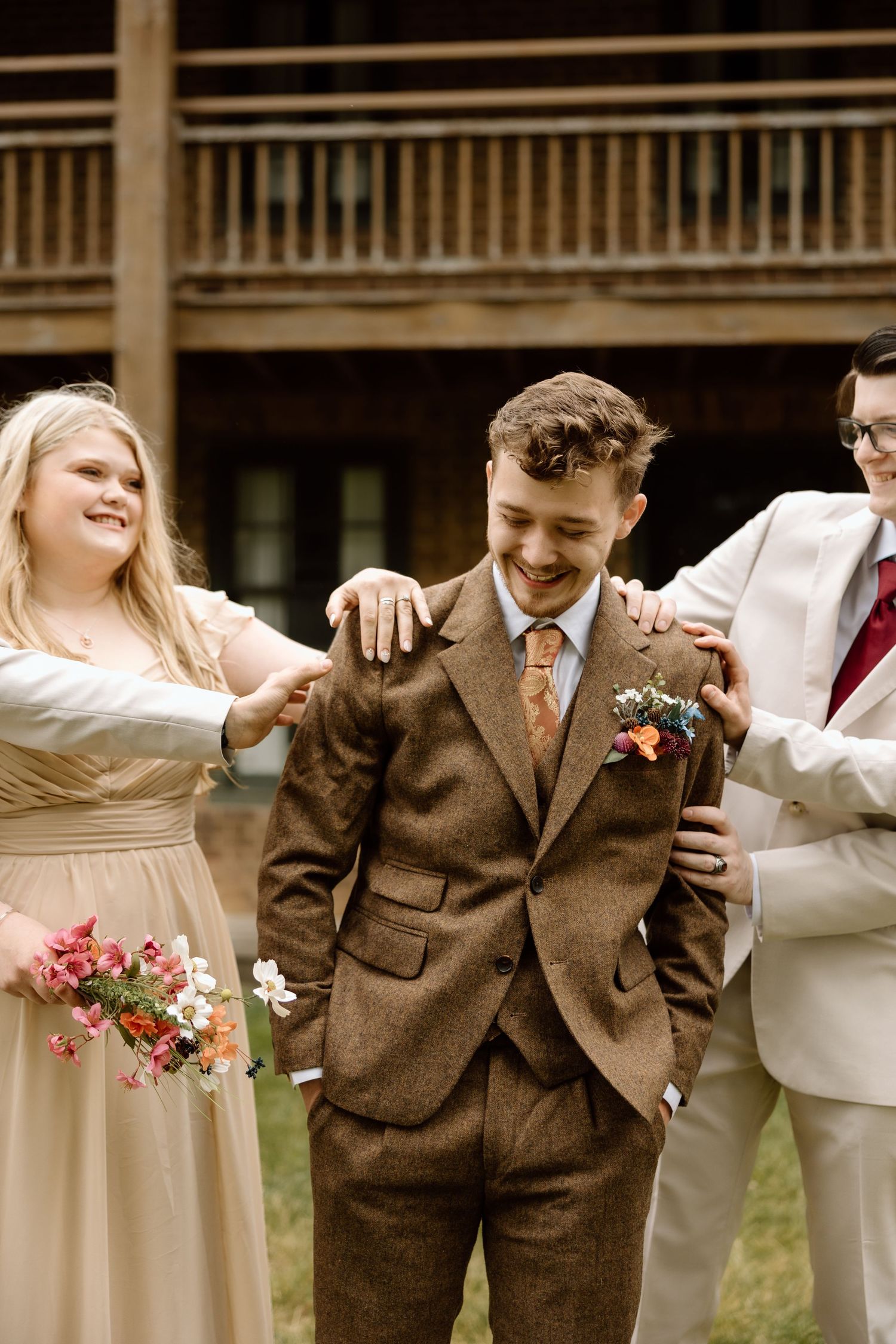 Laughing groom in brown tweed suit being congratulated by bridesmaid and groomsman at outdoor wedding.
