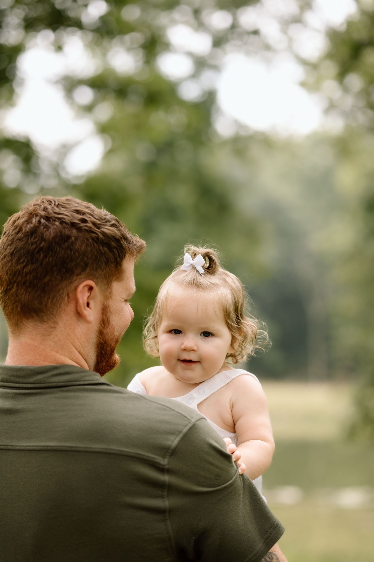 Baby girl with bow in hair looks over fathers shoulder in a lush green park setting.