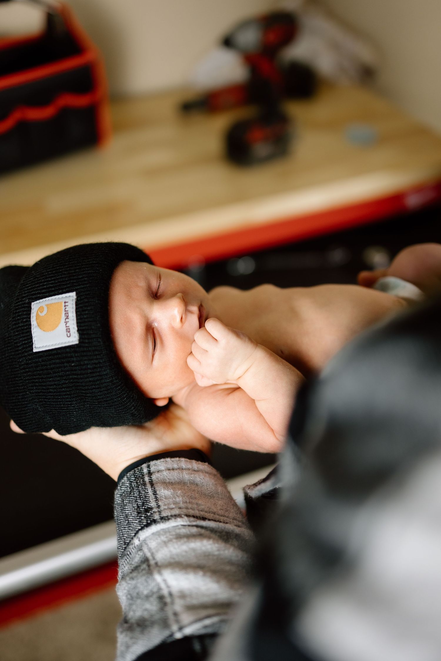 Newborn baby wearing a black Carhartt beanie being cradled in parent hands near a tool workbench.