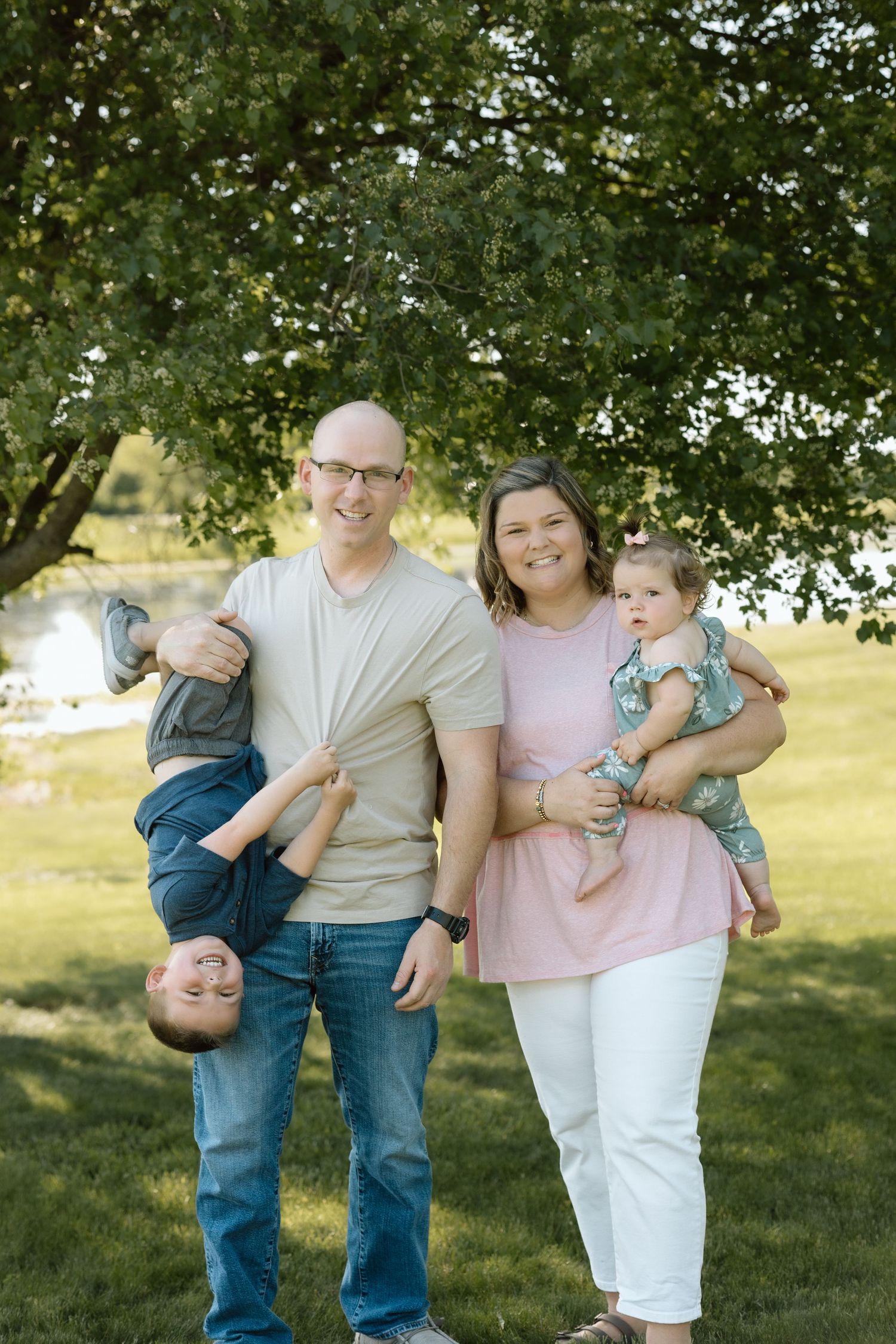 Happy family of four poses outdoors, father holding son upside down while mother cradles baby girl by pond.