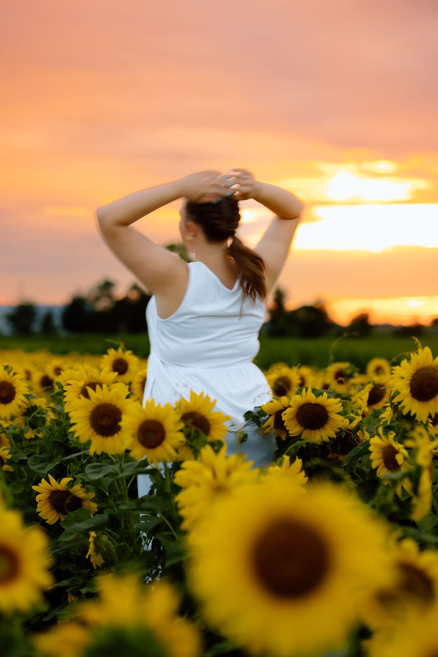 Woman in white dress with hands in hair standing in a sunflower field at colorful sunset.
