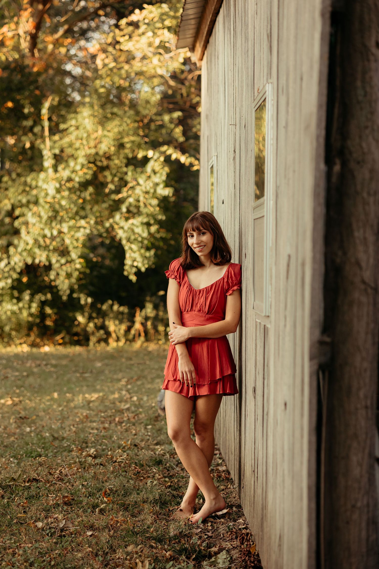 Young woman in red dress leaning against a weathered barn wall at golden hour.
