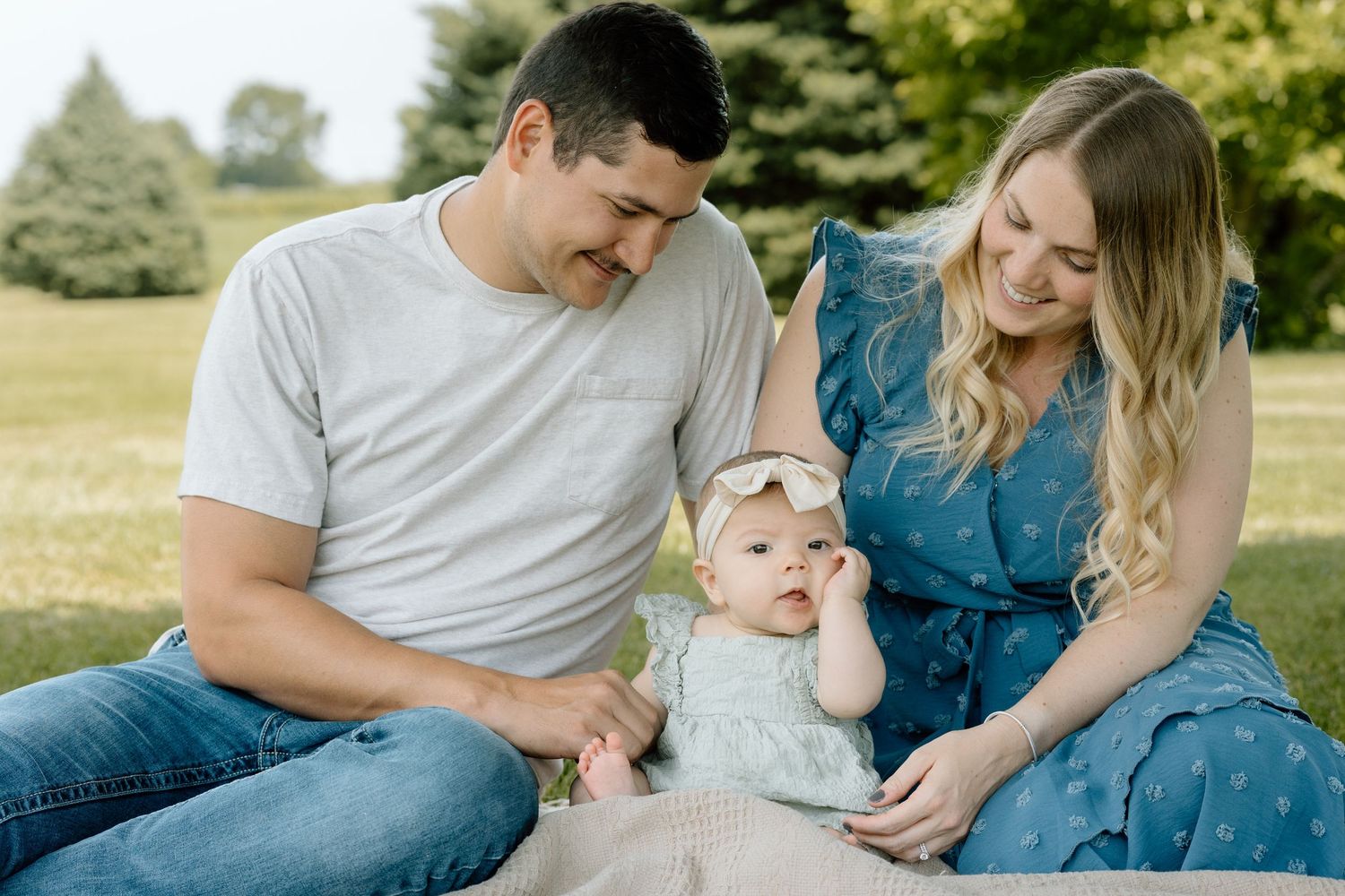 Young family sitting on blanket in green park, parents smiling at baby girl with bow headband.