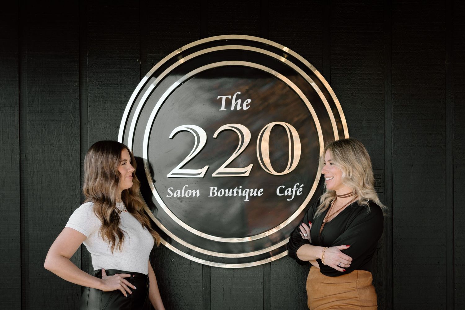 Two women smiling beside The 220 Salon Boutique Cafe circular sign on dark wall.