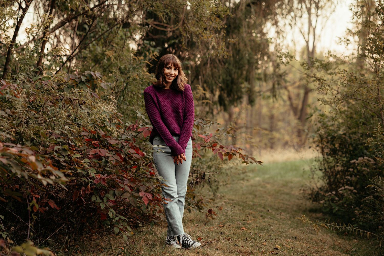 Smiling young woman in burgundy sweater and jeans posing on woodland path surrounded by fall foliage.