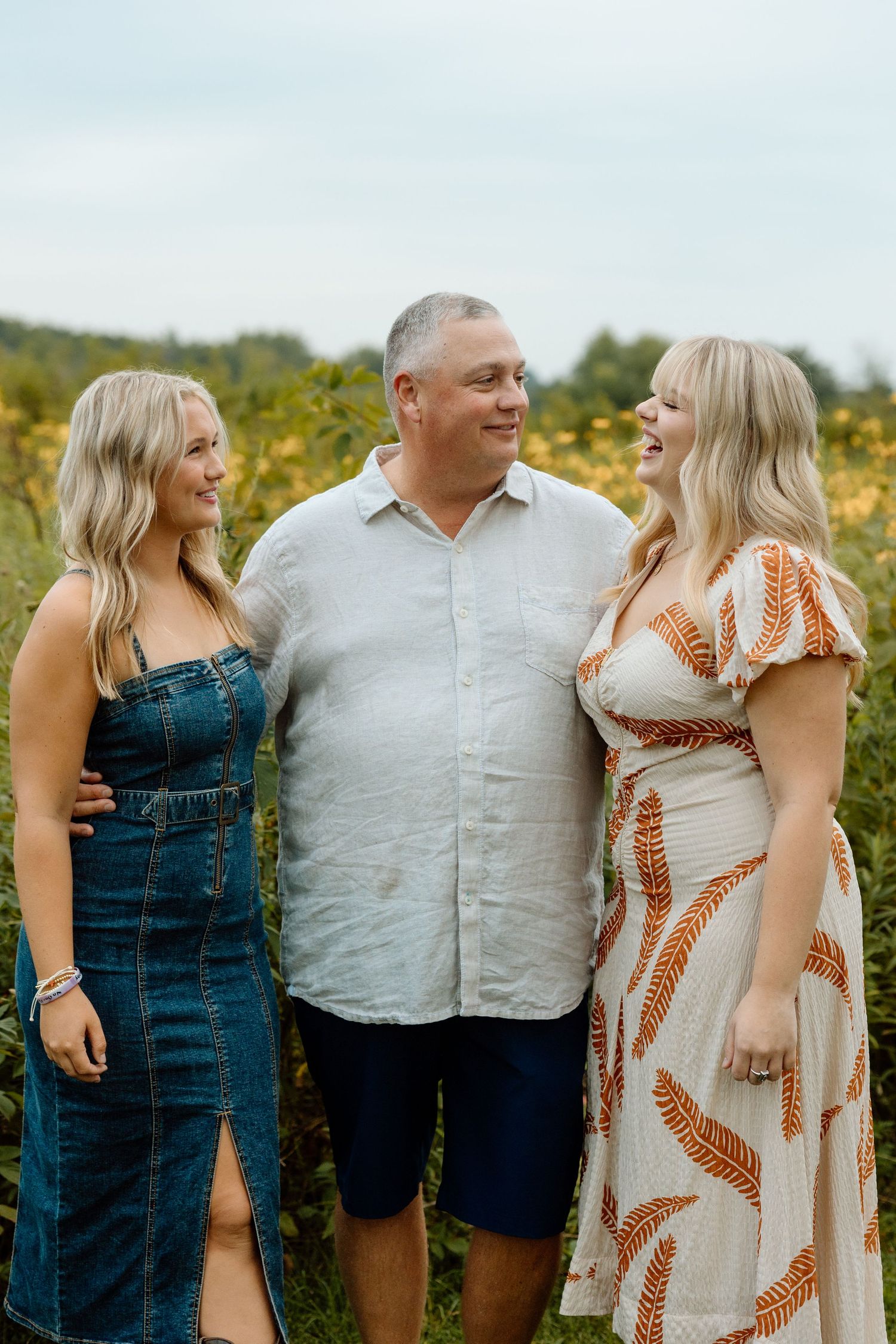 Father and two blonde daughters laughing together in a lush yellow wildflower field outdoors.