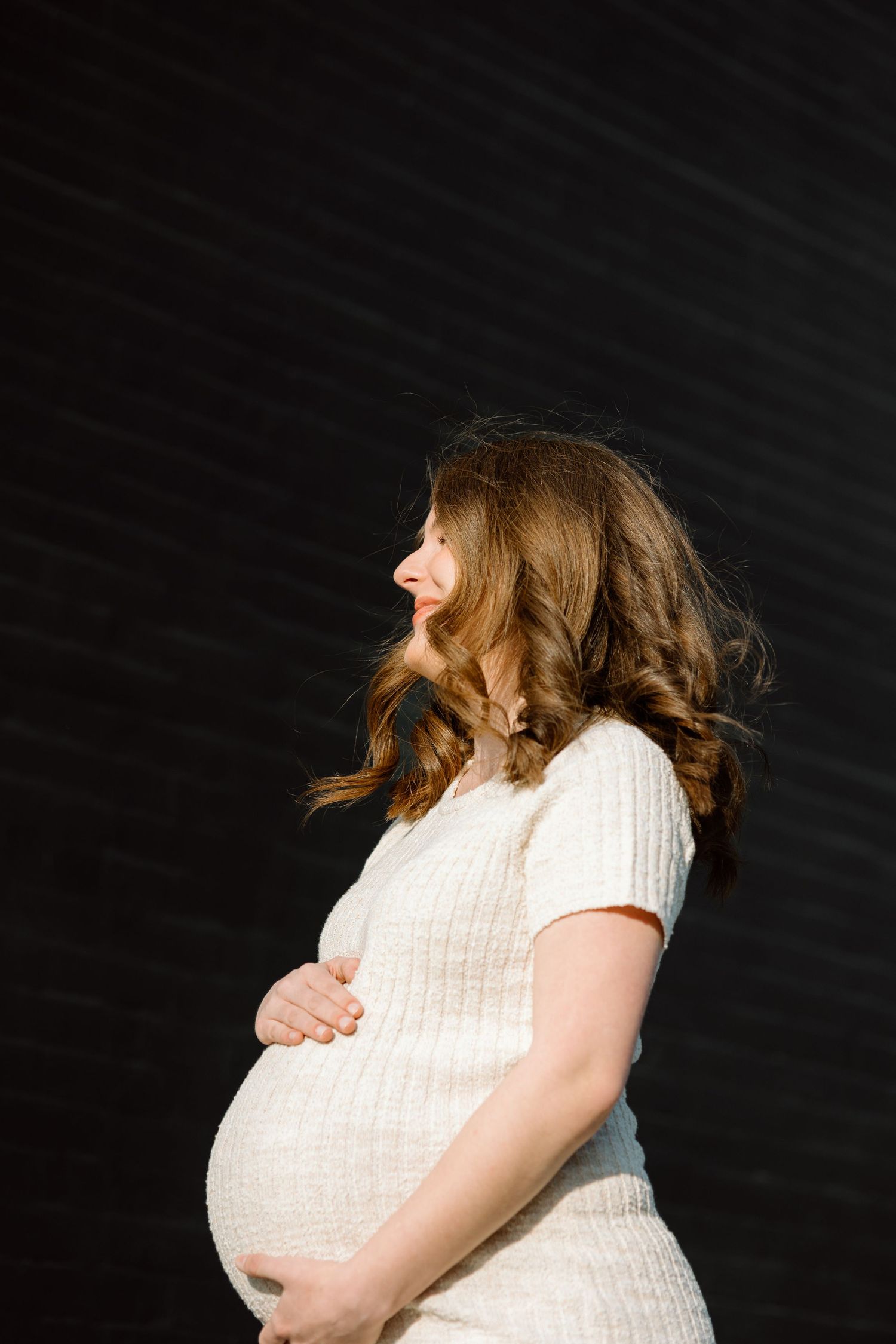 Pregnant woman in white knit dress holding her baby bump while standing in profile against dark wall.