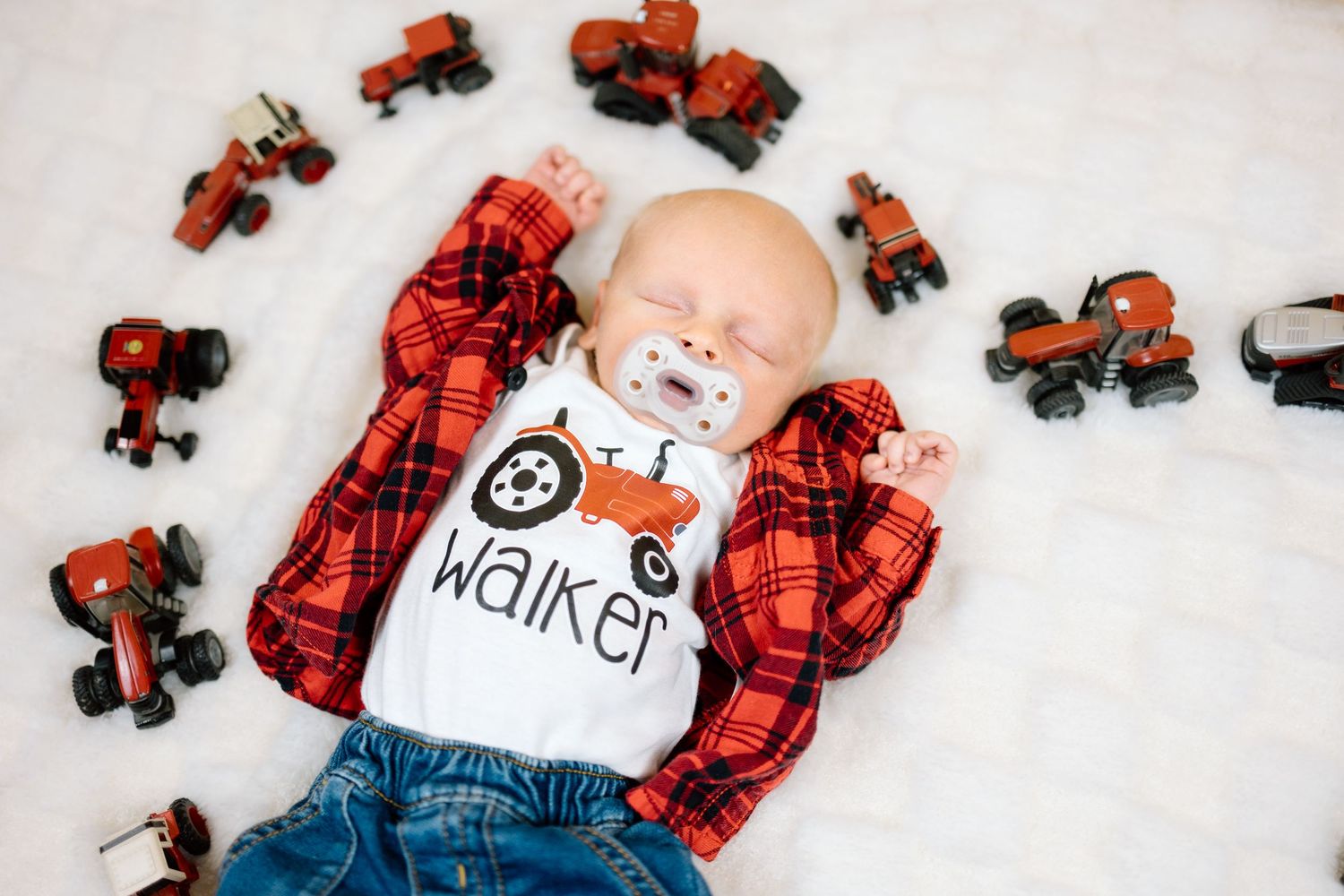 Sleeping baby named Walker in red plaid flannel shirt and jeans surrounded by orange toy tractors on white fluffy surface.