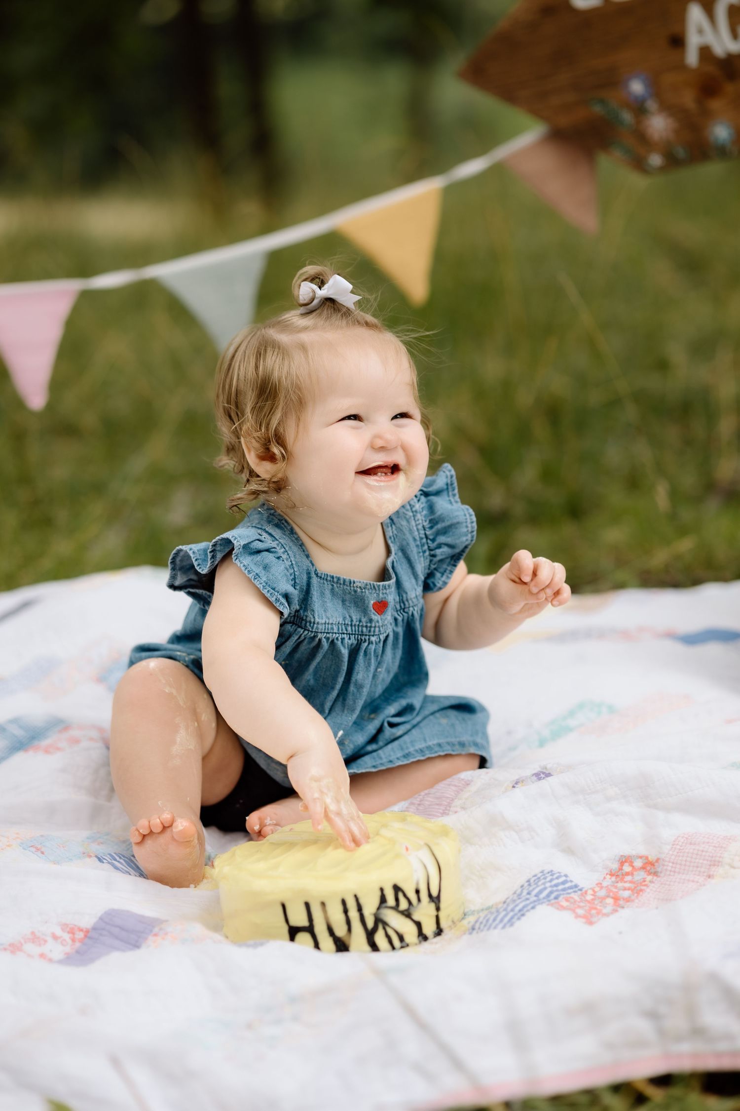 Laughing baby girl in denim dress smashing her first birthday cake outdoors on a quilt.