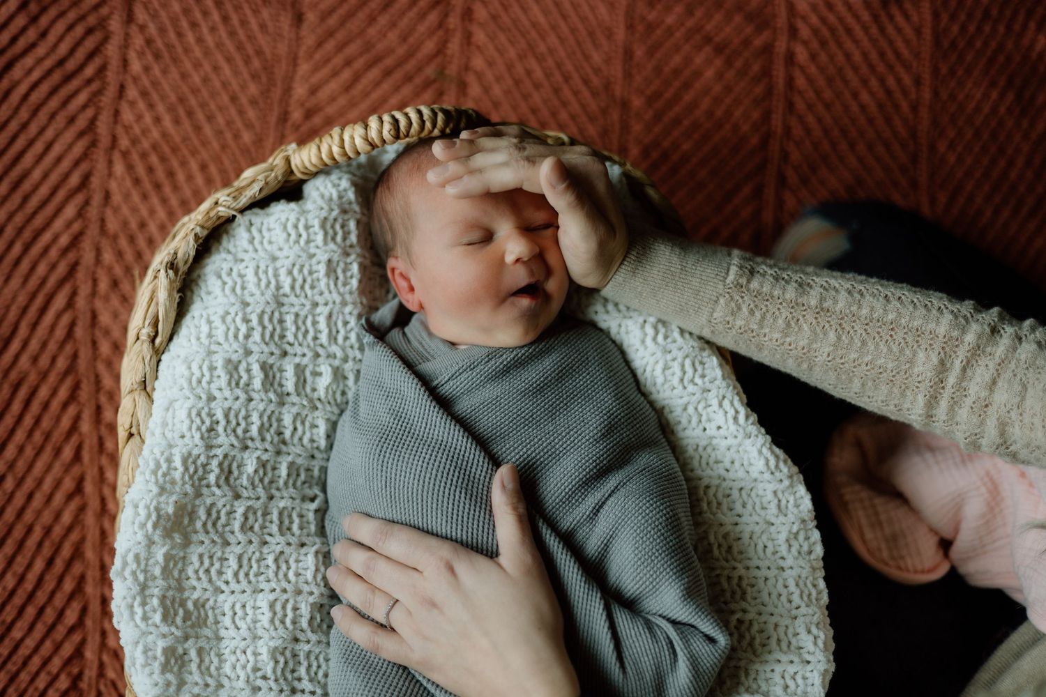 Parent gently touches sleeping newborn baby swaddled in gray wrap inside wicker basket.