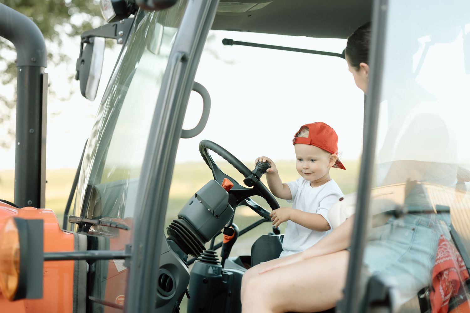 Young child in red cap pretending to drive a red tractor with an adult beside him.