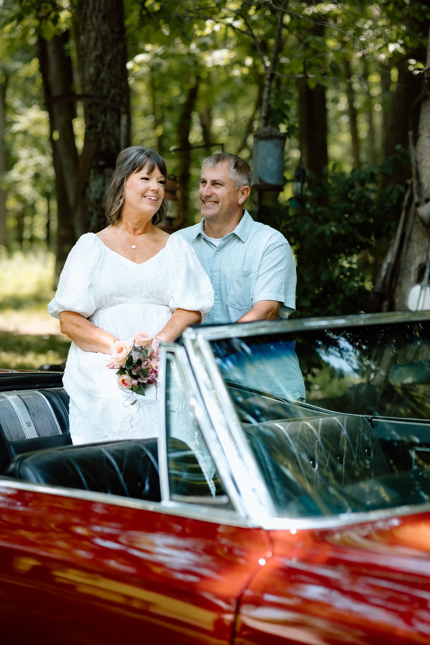 Smiling bride holding bouquet and groom standing beside red vintage convertible in wooded setting.