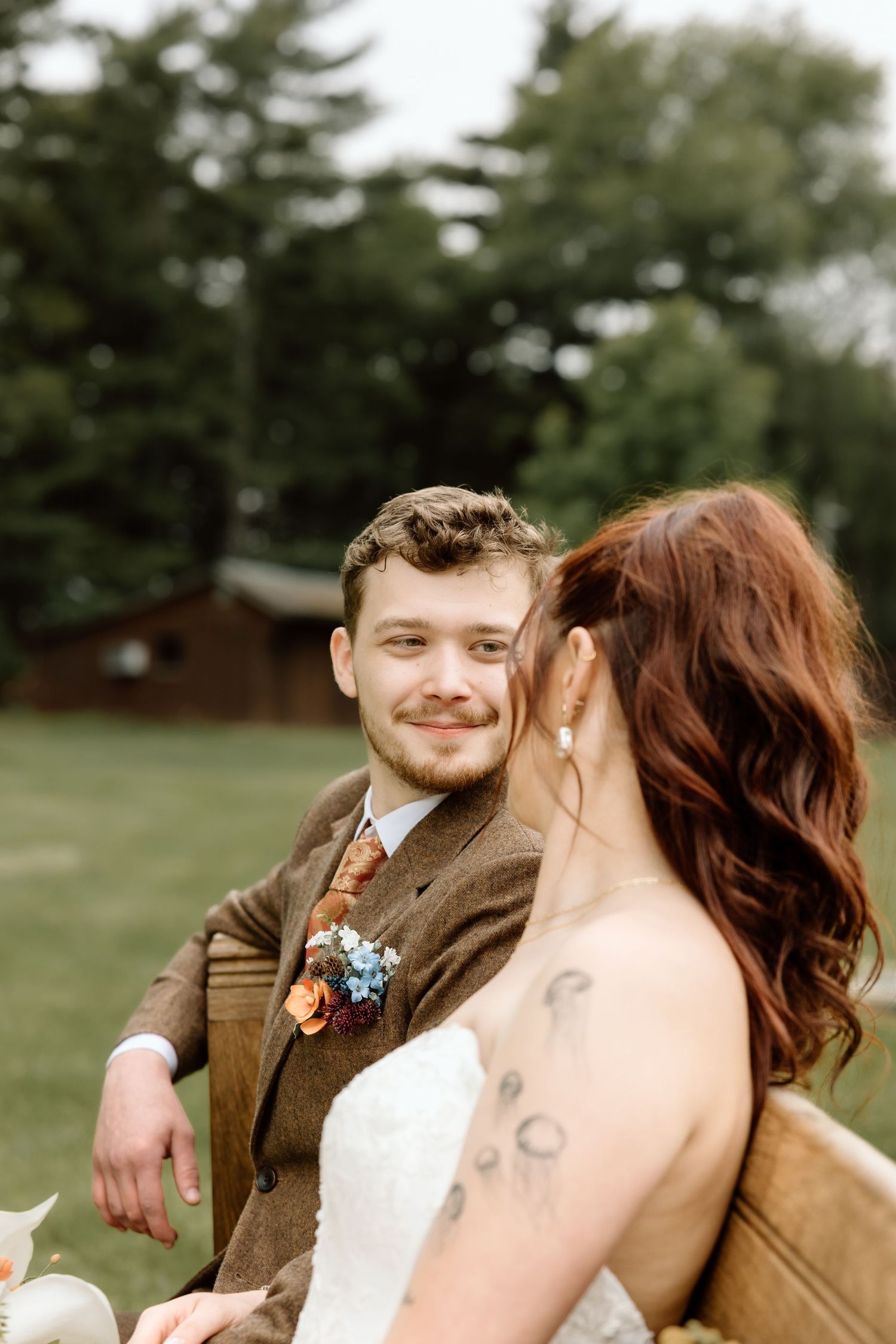 Smiling groom in brown tweed suit gazing at tattooed bride with red hair during outdoor wedding portraits.
