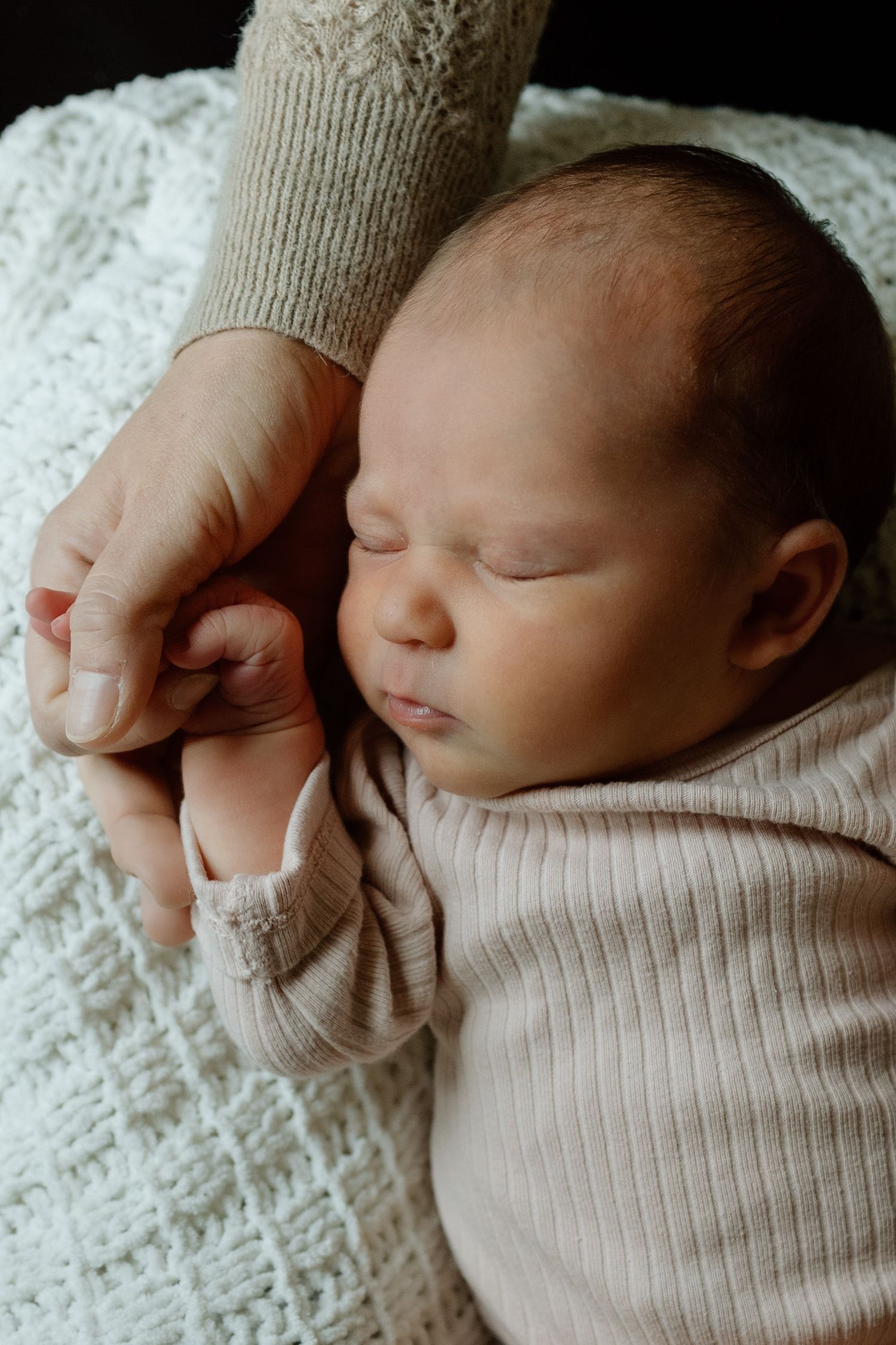 Sleeping newborn baby in pink onesie gently holding parent's hand on white knit blanket.