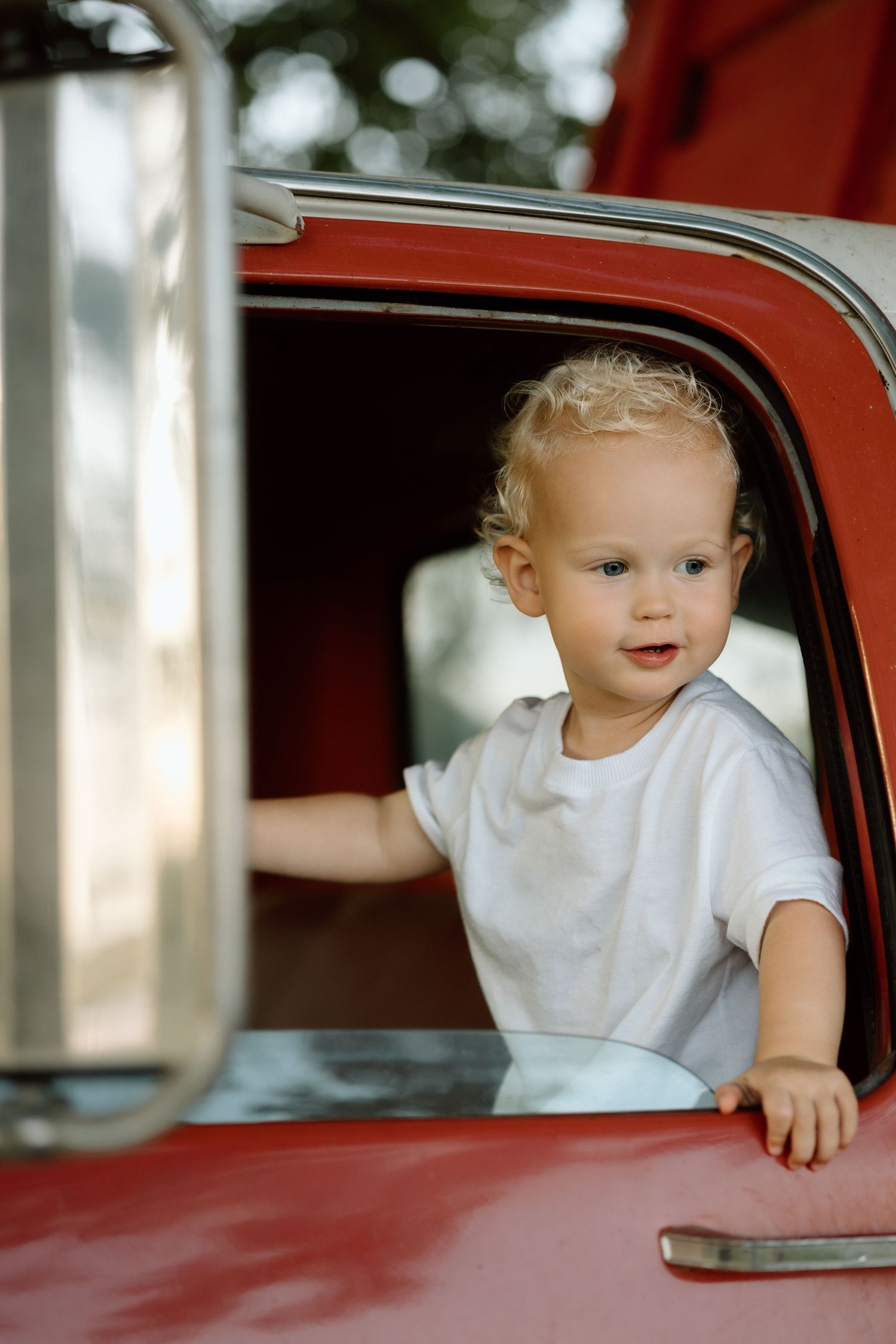 Adorable blond toddler in white shirt leans out open window of classic red truck.