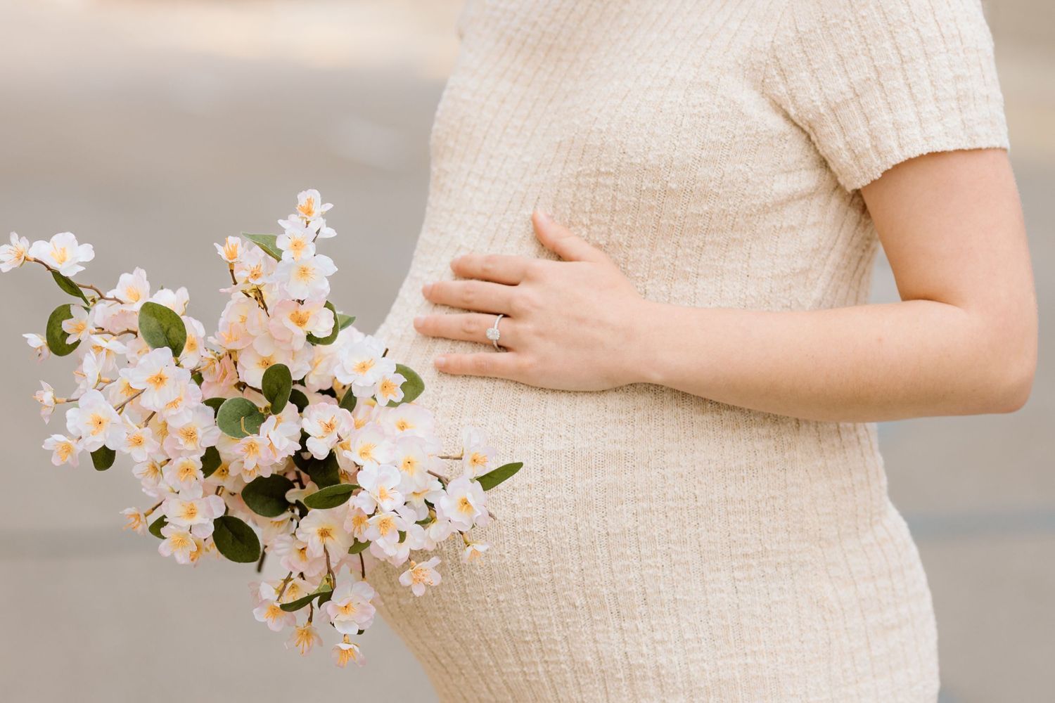 Pregnant woman in cream knit dress holding white flowers against her baby bump during maternity photoshoot.