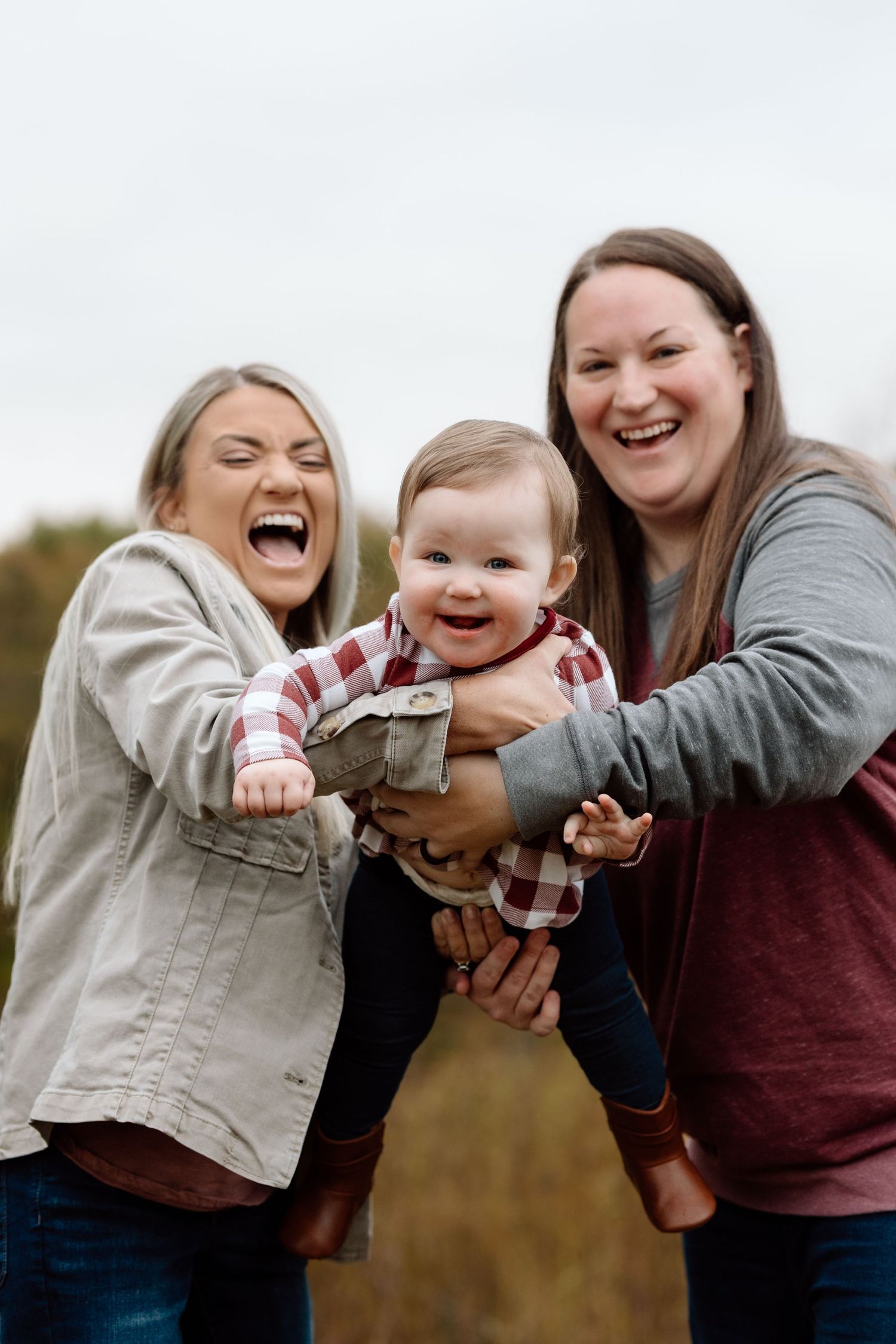 Two laughing women holding a happy baby in a red plaid shirt during an outdoor fall photo session.