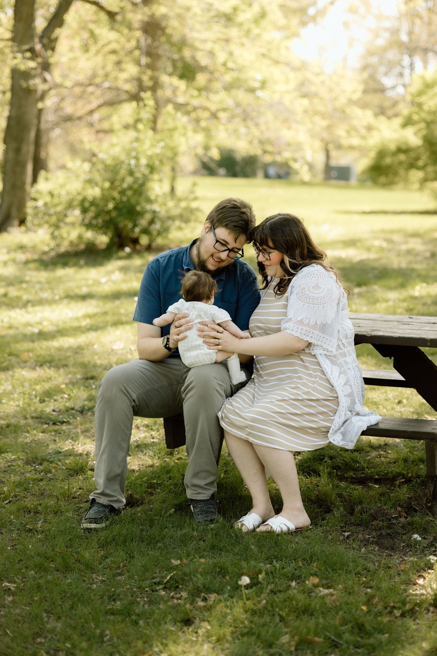 New parents sit close together on park bench, tenderly cradling their newborn baby in dappled sunlight.