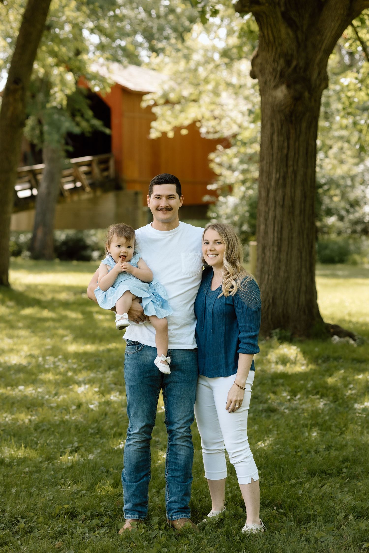 Young family of three poses outdoors with parents holding baby girl in blue dress near large trees and red barn.