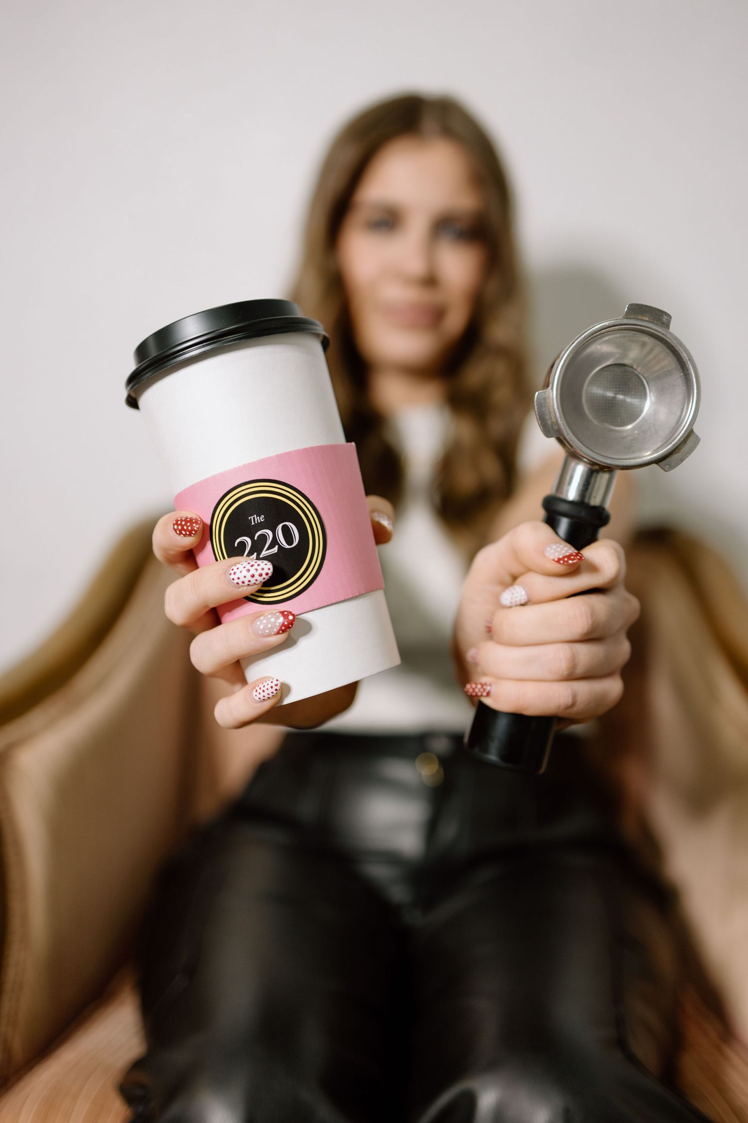 Blonde woman holding The 220 branded pink coffee cup and espresso portafilter while seated in chair.
