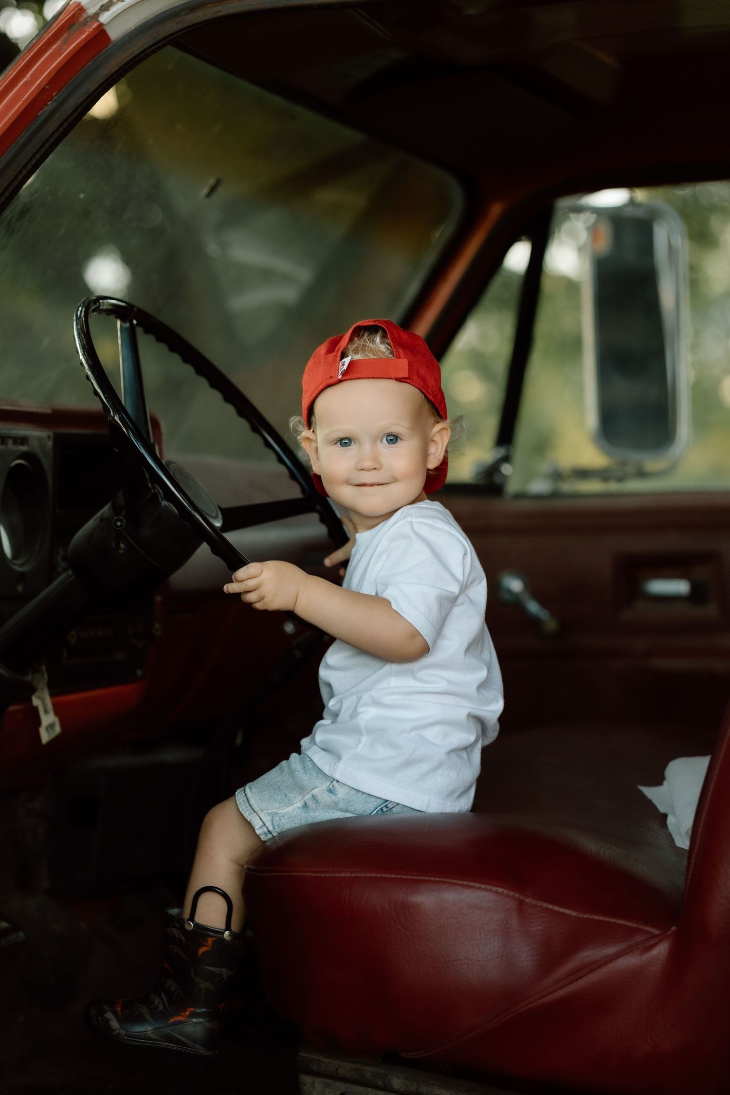 Smiling toddler boy in red baseball cap sits behind steering wheel of classic red truck.