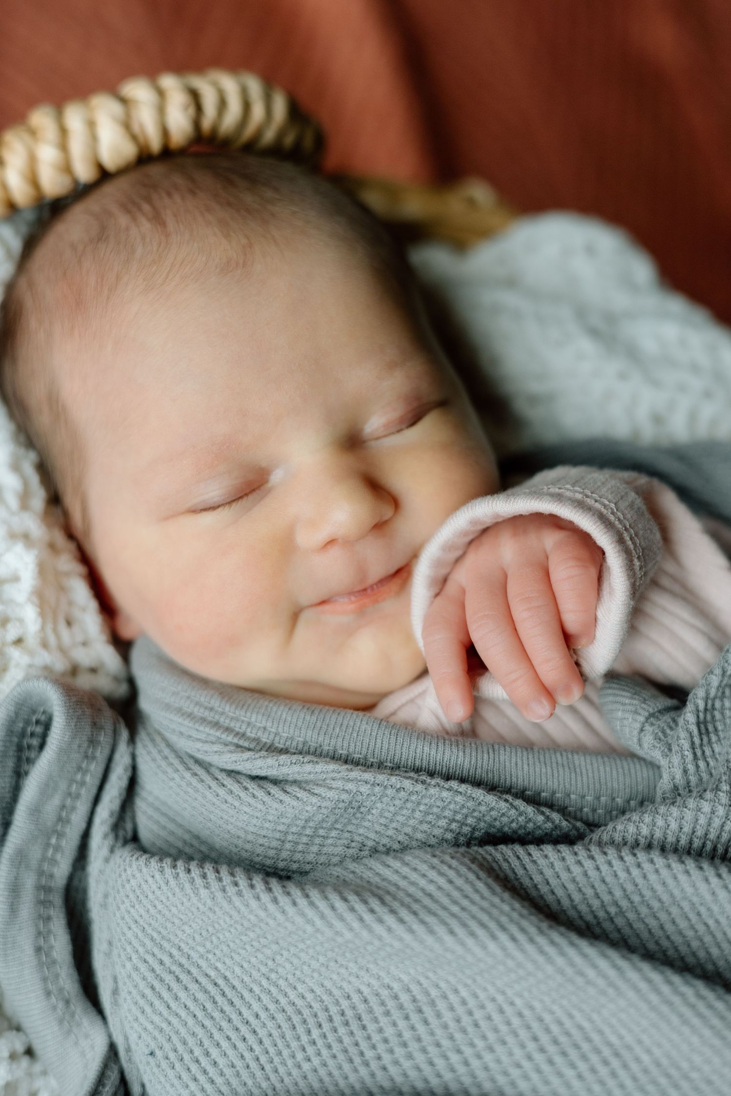 Smiling sleeping newborn baby swaddled in gray blanket with tiny hand visible in wicker basket.