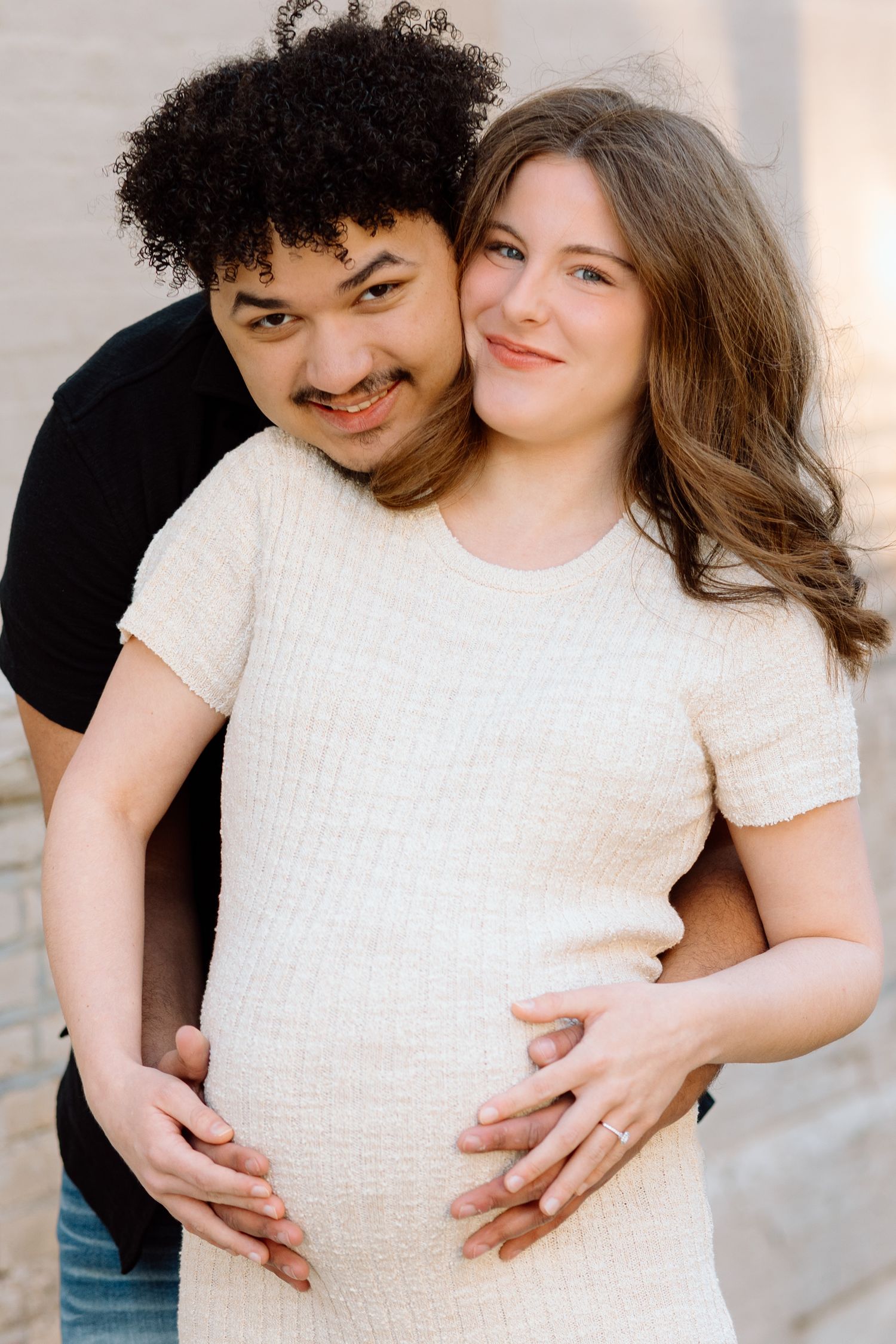 Interracial couple maternity photo, man hugging pregnant woman in cream dress from behind, both smiling warmly.