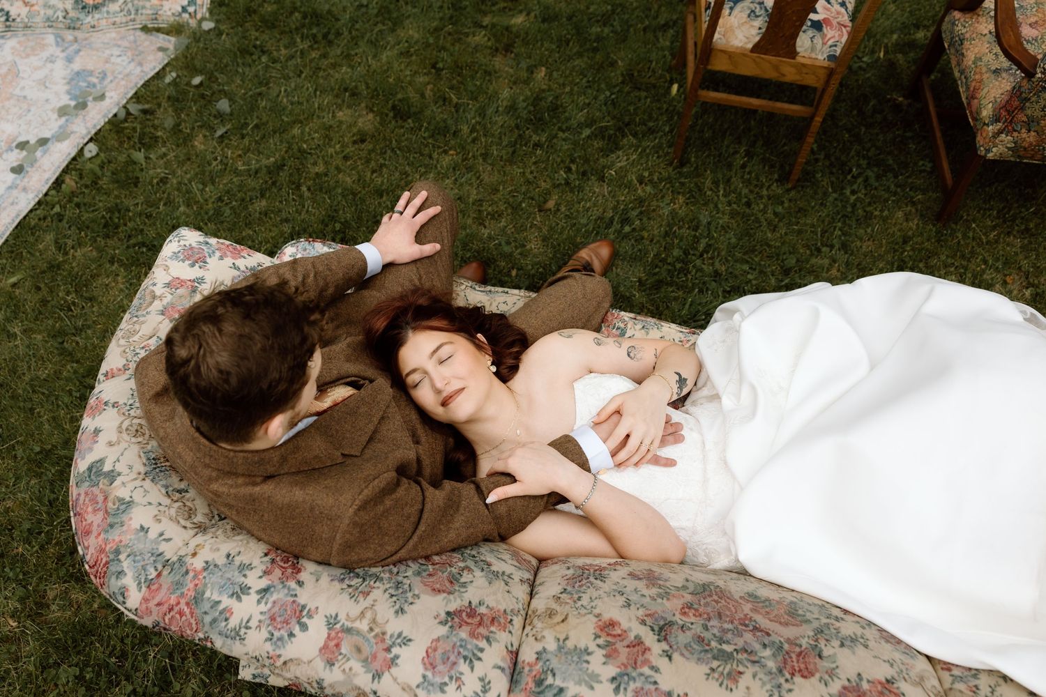 Bride and groom lying on a floral mattress on green grass during an outdoor wedding photo session.