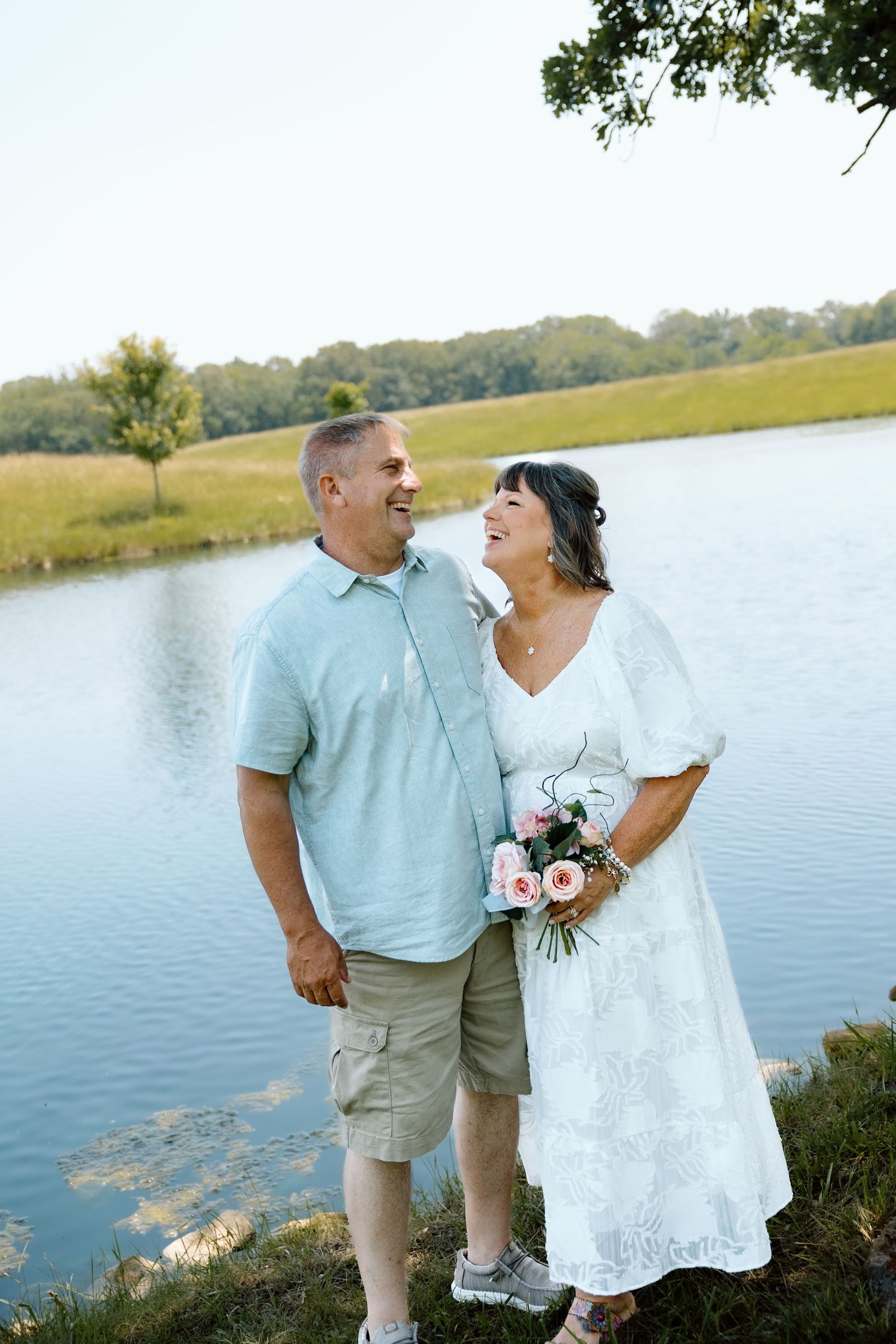 Smiling couple in wedding attire standing by a serene lake, bride holding pink roses bouquet.