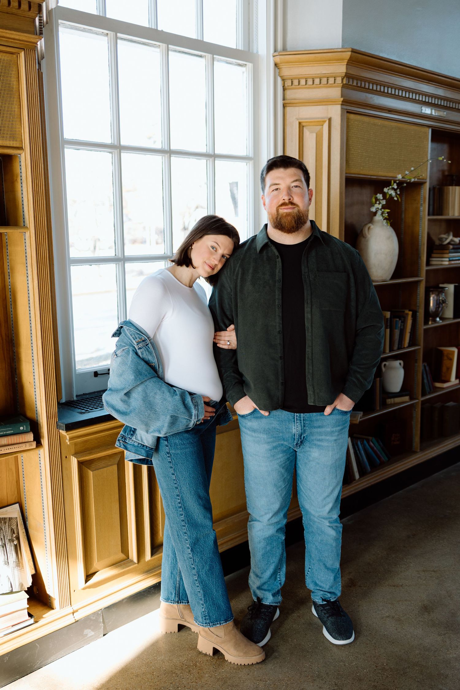 Couple stands by window in cozy interior, woman leans on mans shoulder during maternity session.