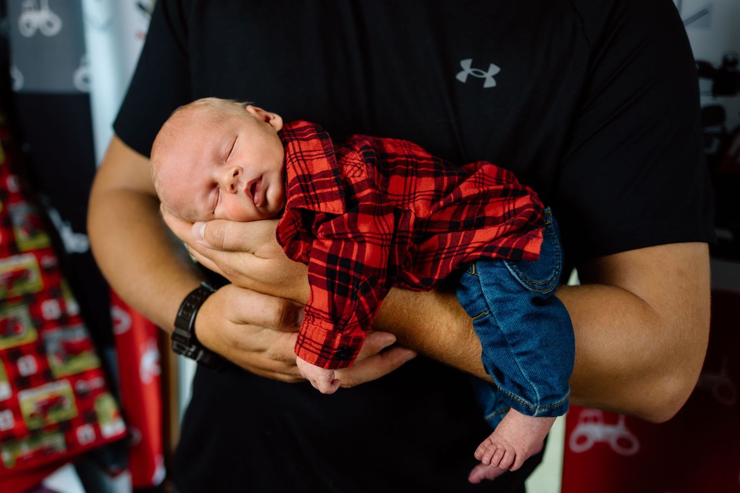 Father in black shirt cradling sleeping newborn dressed in red plaid shirt and jeans.