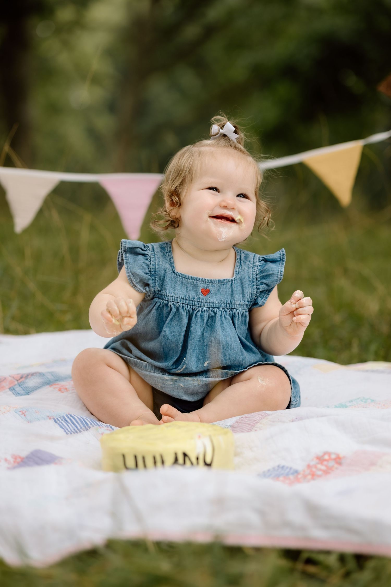 Happy baby girl in denim dress laughing beside yellow smash cake on quilt with bunting flags.