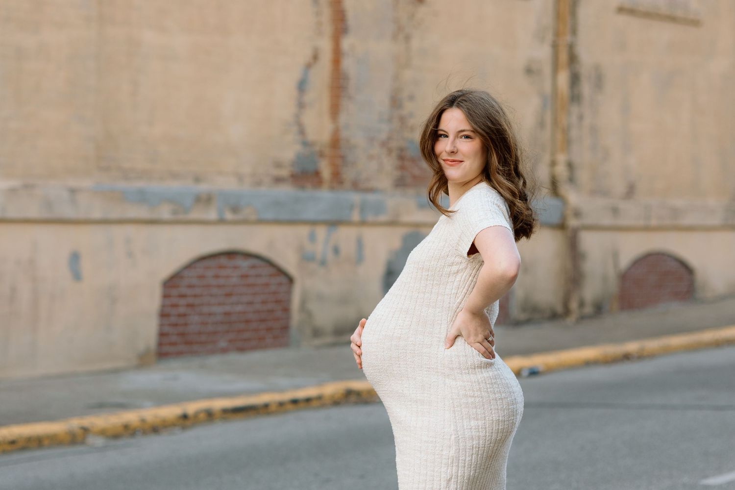 Beautiful pregnant woman in white knit dress posing on a street with vintage brick building backdrop.