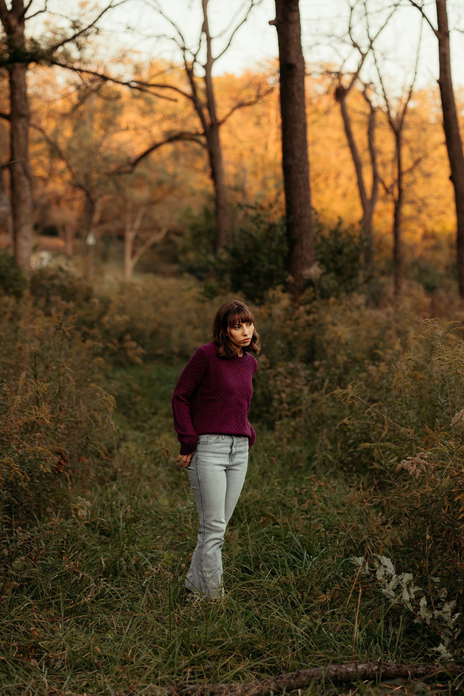 Young woman in purple sweater stands in tall grass with colorful autumn trees in background.