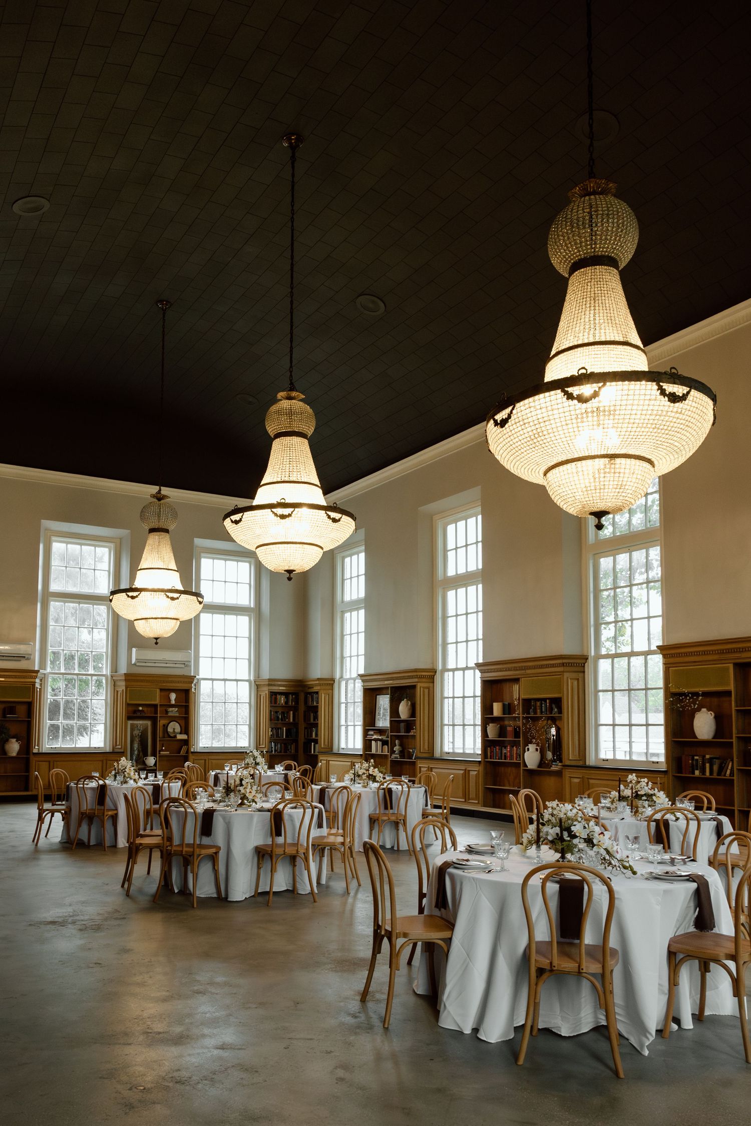 Grand event hall with crystal chandeliers, white-draped round tables, and tall arched windows.