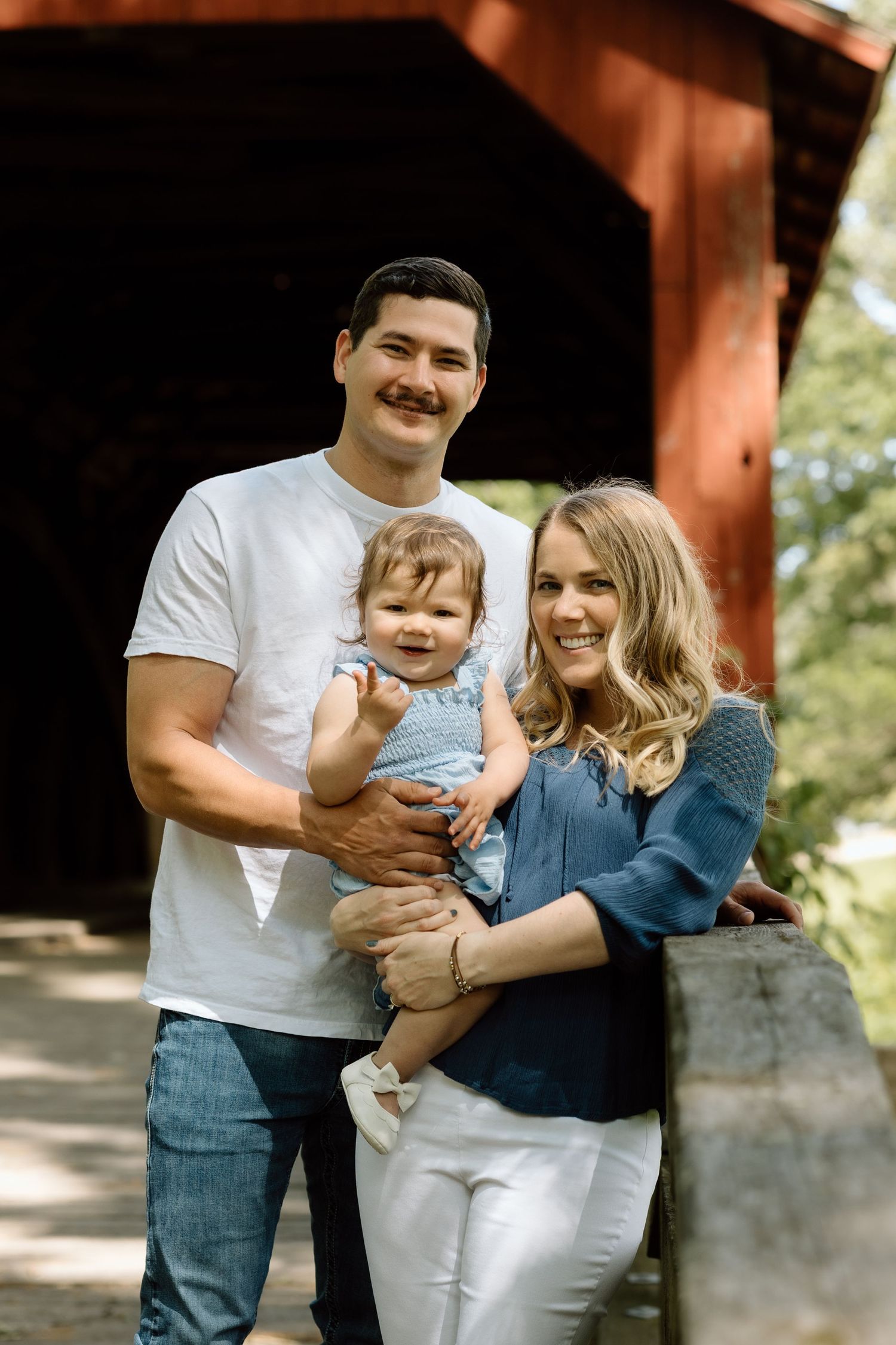 Young family poses with baby daughter in blue dress in front of a red covered bridge during outdoor photo session.